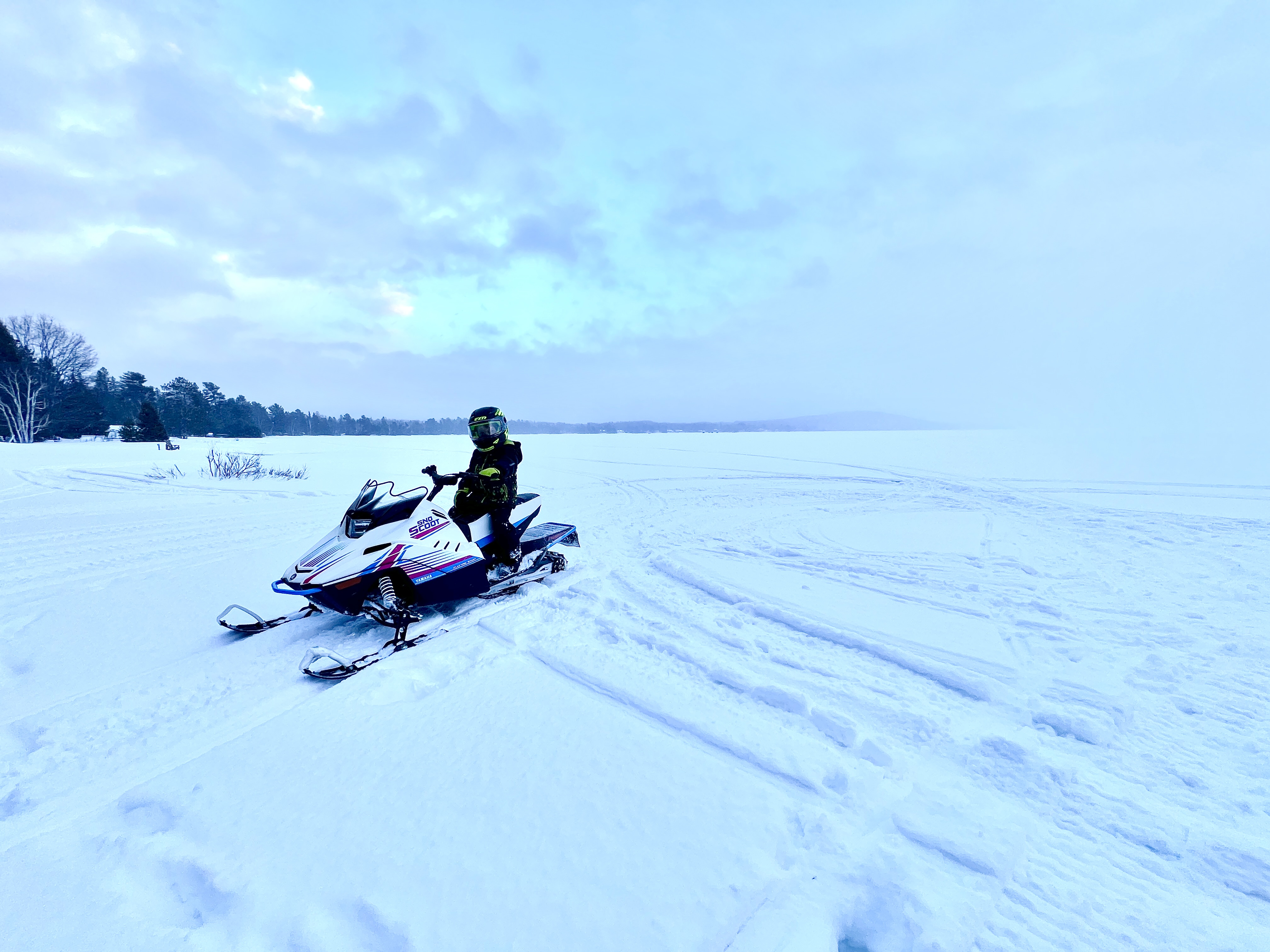 A boy on a Snoscoot snowmobile in an open, snow-covered field with forest in the horizon under a soft blue, clouded winter sky.