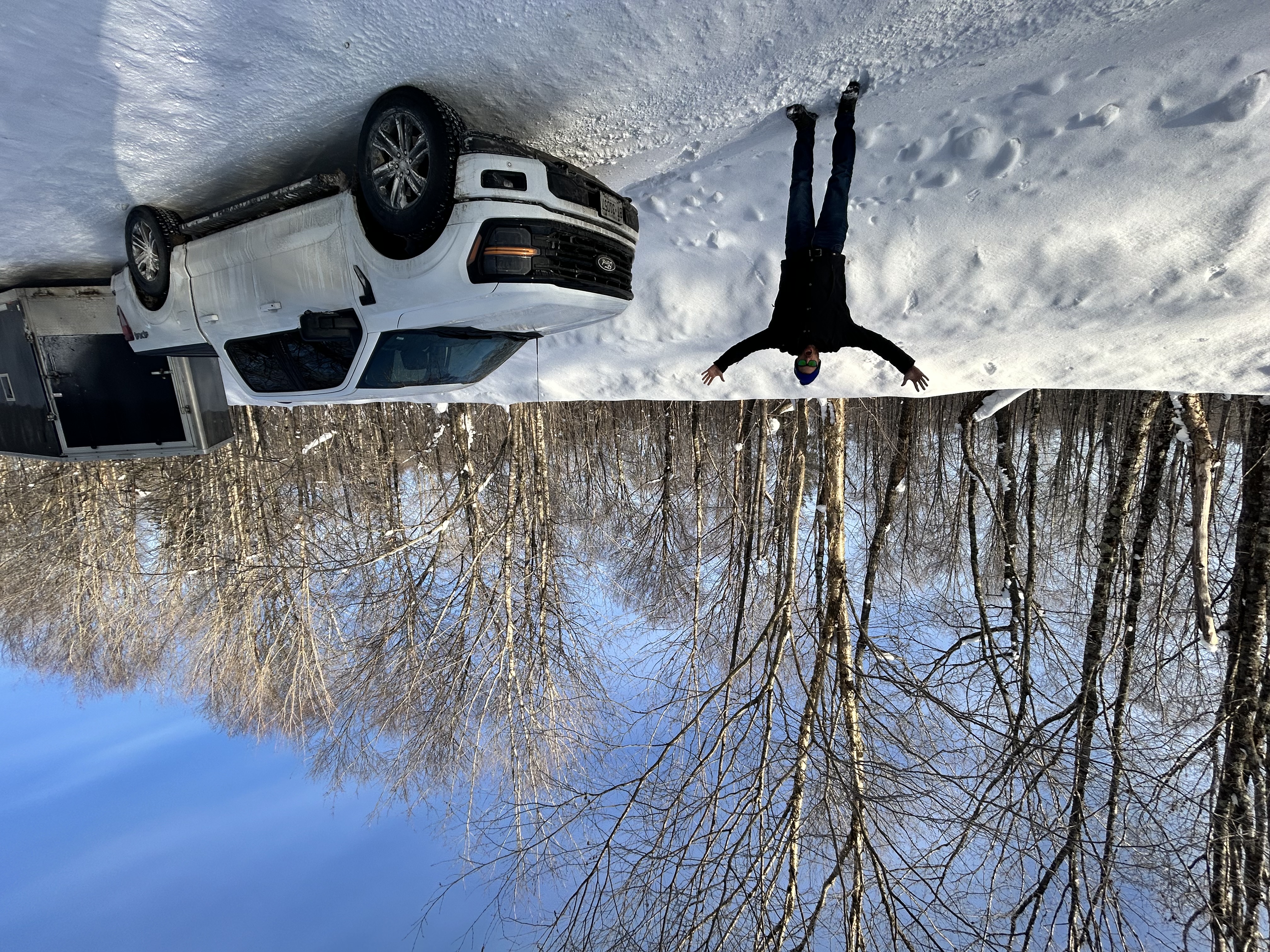 A man standing next to his truck and trailer, with his arms raised excitedly as he gestures to the deep snow covering the snowmobile trail and forest all around.