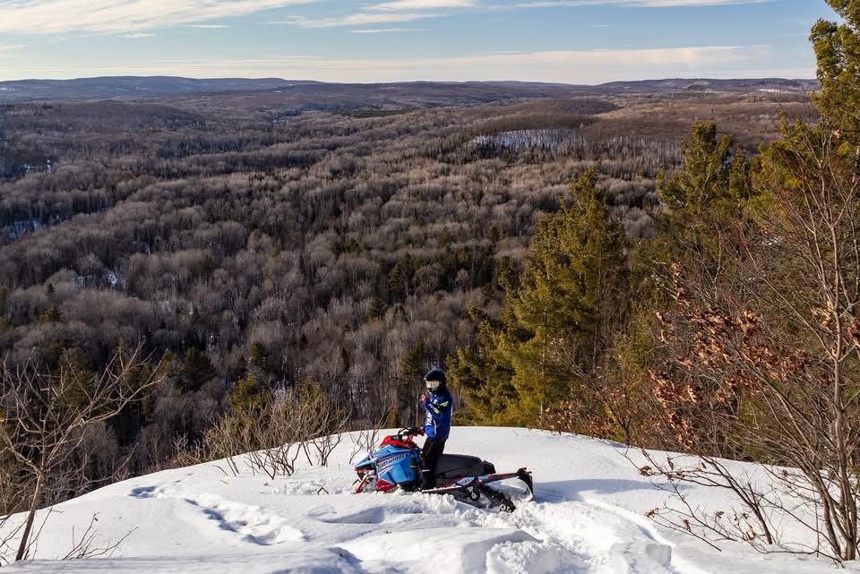A snowmobiler standing astride their machine at the top of a snowy hill, looking out over a broad view of a wide, forested valley far below. 