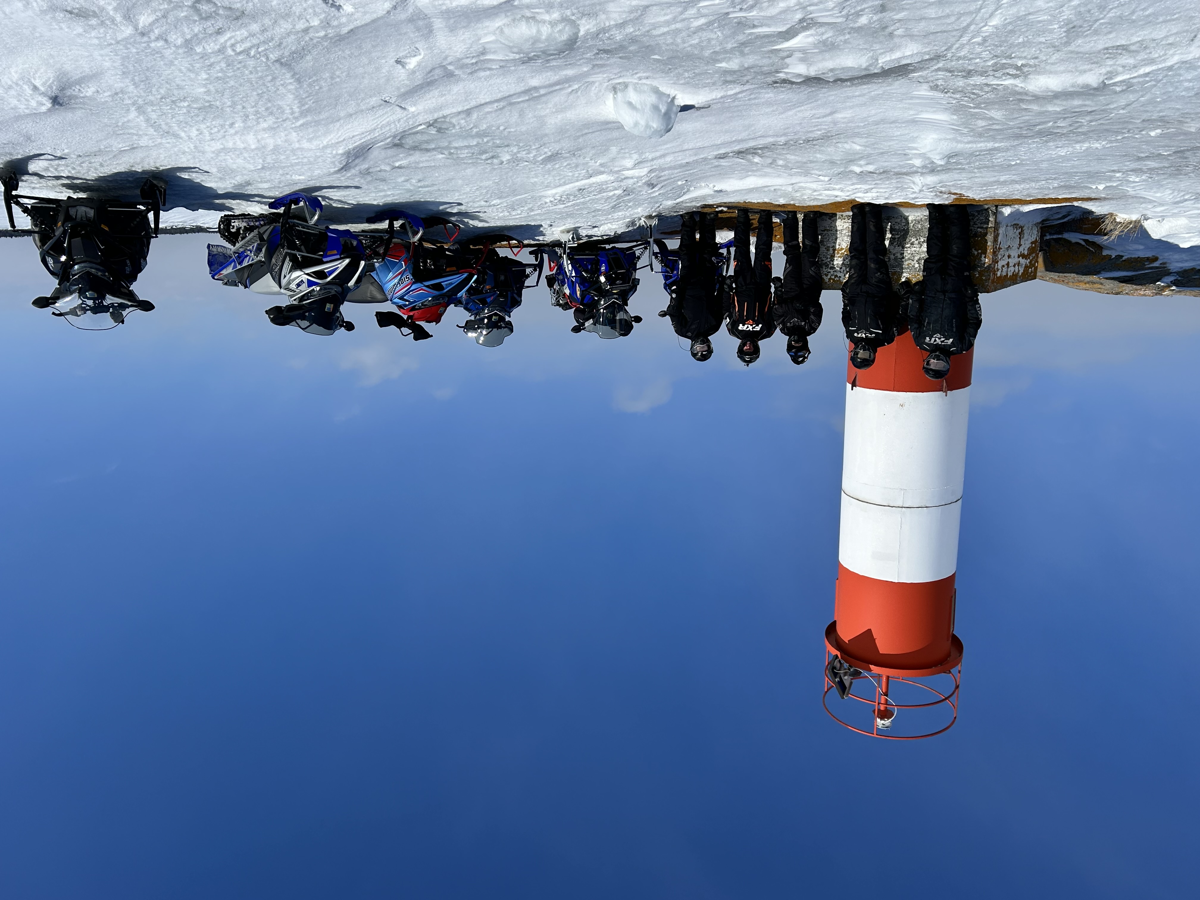 A group of snowmobilers standing next to a red and white Georgian Bay lighthouse on a snowy ridge in front of a vivid, clear blue winter sky.