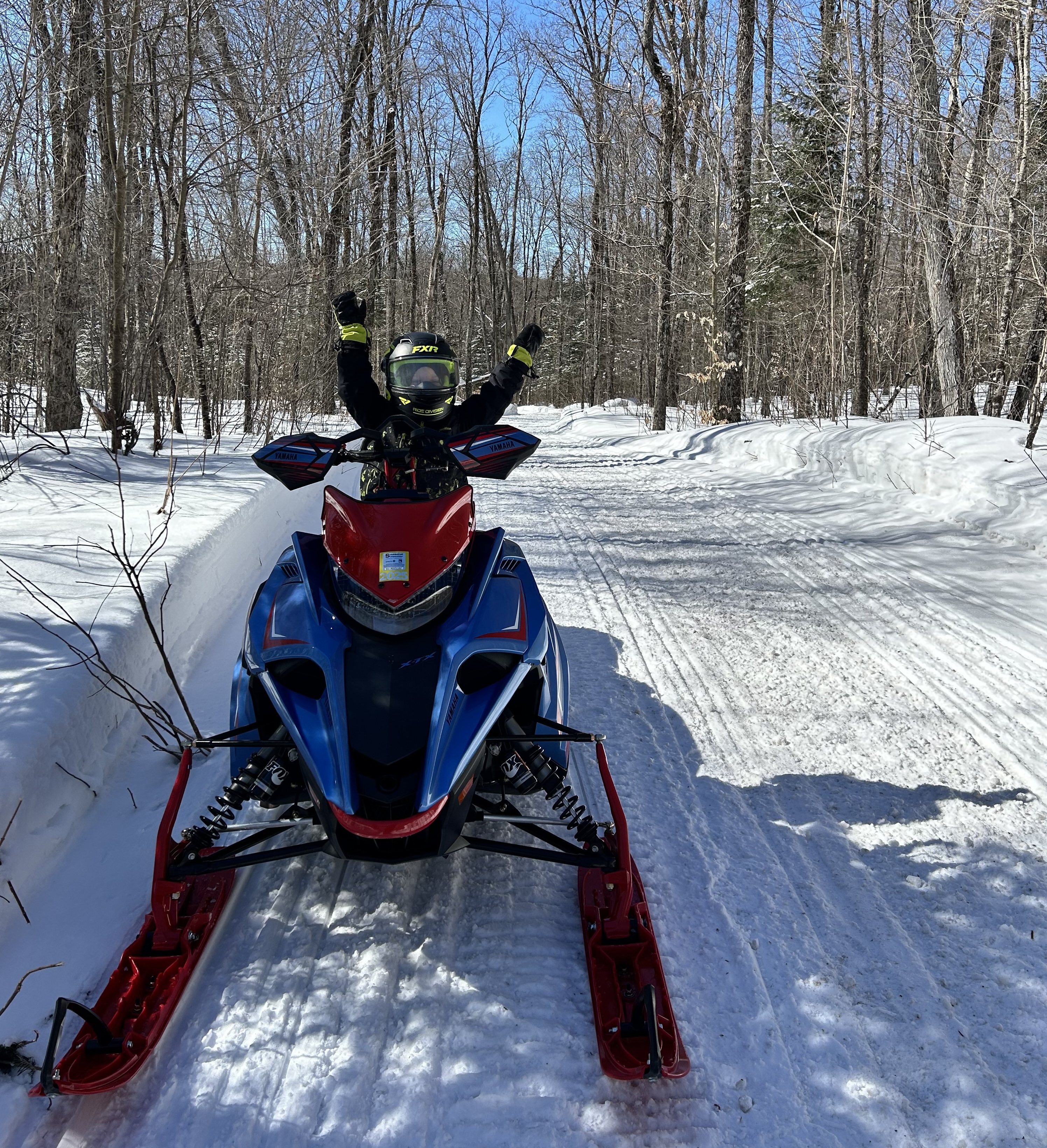 a small boy in snowmobile gear sits on a snowmobile on a snowy forest trail, cheering with arms raised over his head.