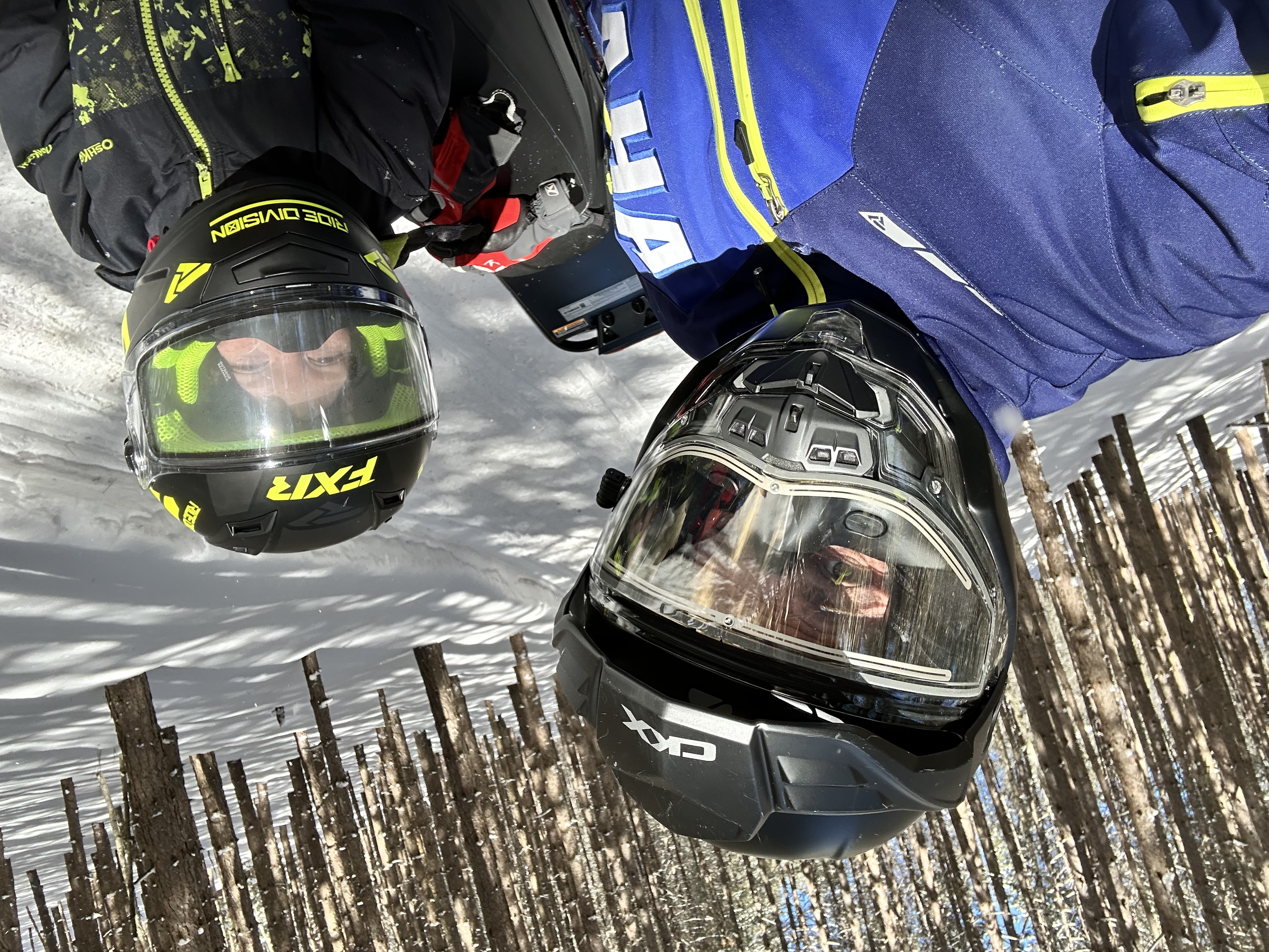 A father and son in snowmobile helmets posing on a forested snowmobile trail.
