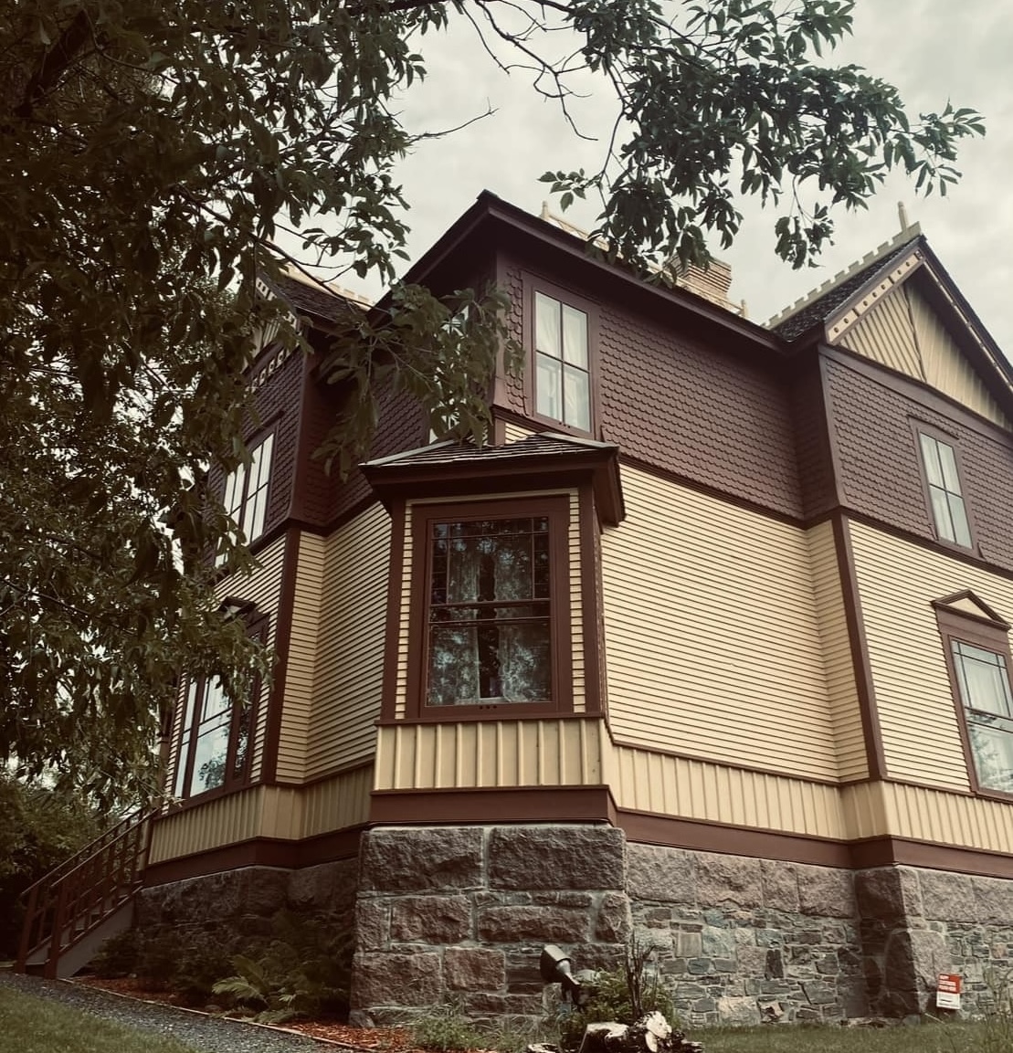a low angle of the Mather Walls Teahouse, a stately and looming Queen Anne style house with stone foundation and tall windows with tree branches hanging overhead.