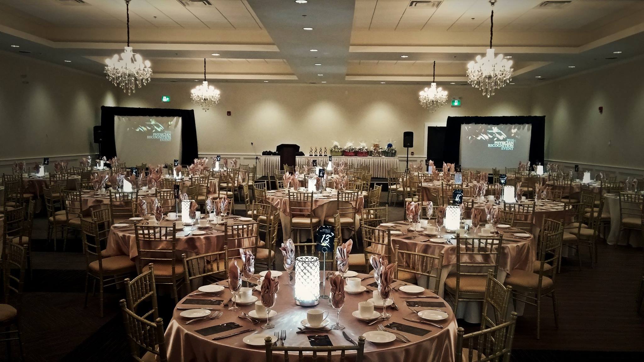 round tables lit by glowing lanterns elegantly set for dinner with white china and pink table linens. Crystal chandeliers glow overhead and viewing screens stand in each corner. 