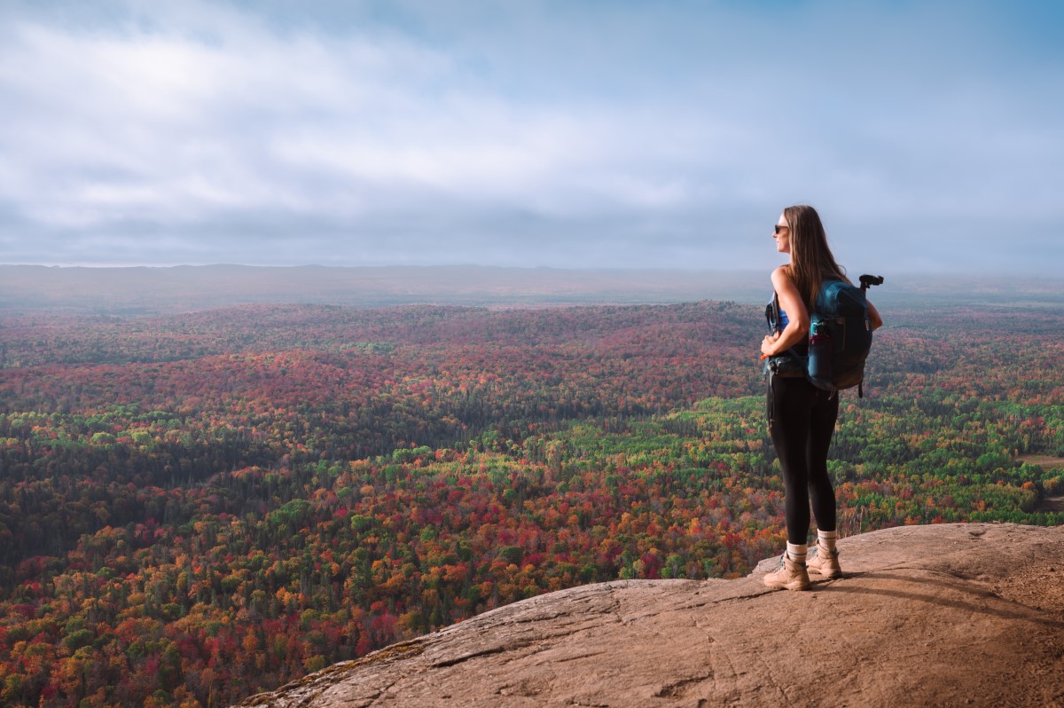 Robertson Cliffs, Sault Ste. Marie; a hiker wearing a backpack stands on a high rock lookout, gazing out over the wide valley full of red and green autumn forest far below.