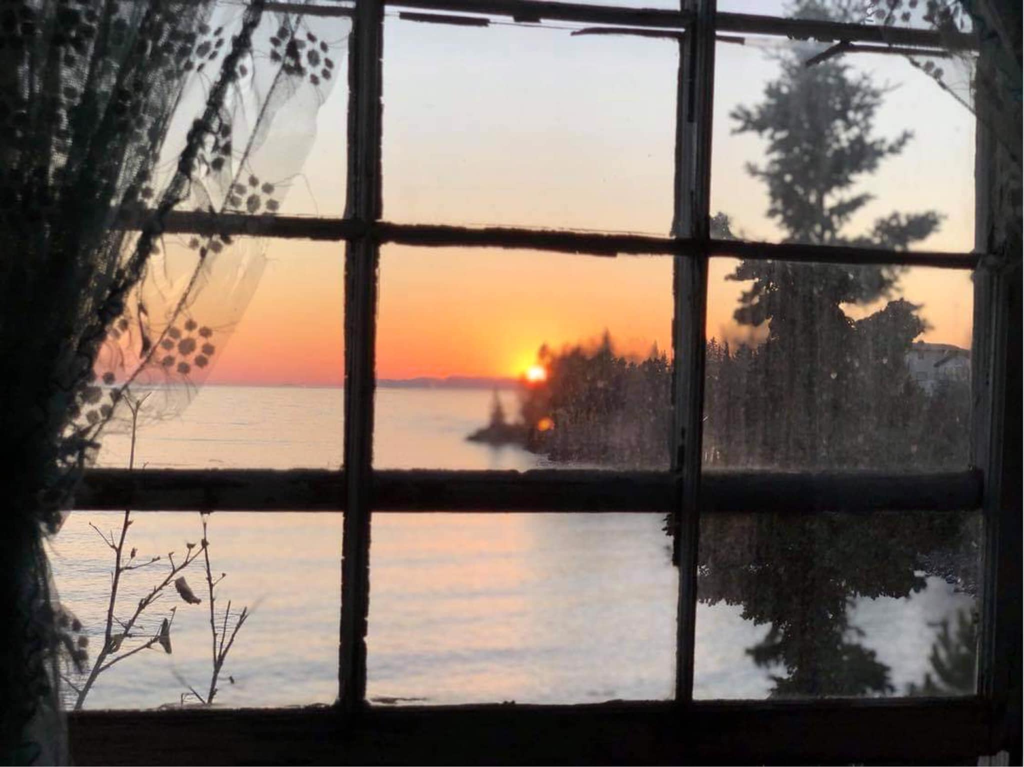 an orange sunrise over the silvery bay surrounded by forest,  seen from inside the Silver Islet General Store through a waving antique paned window with chipping paint.