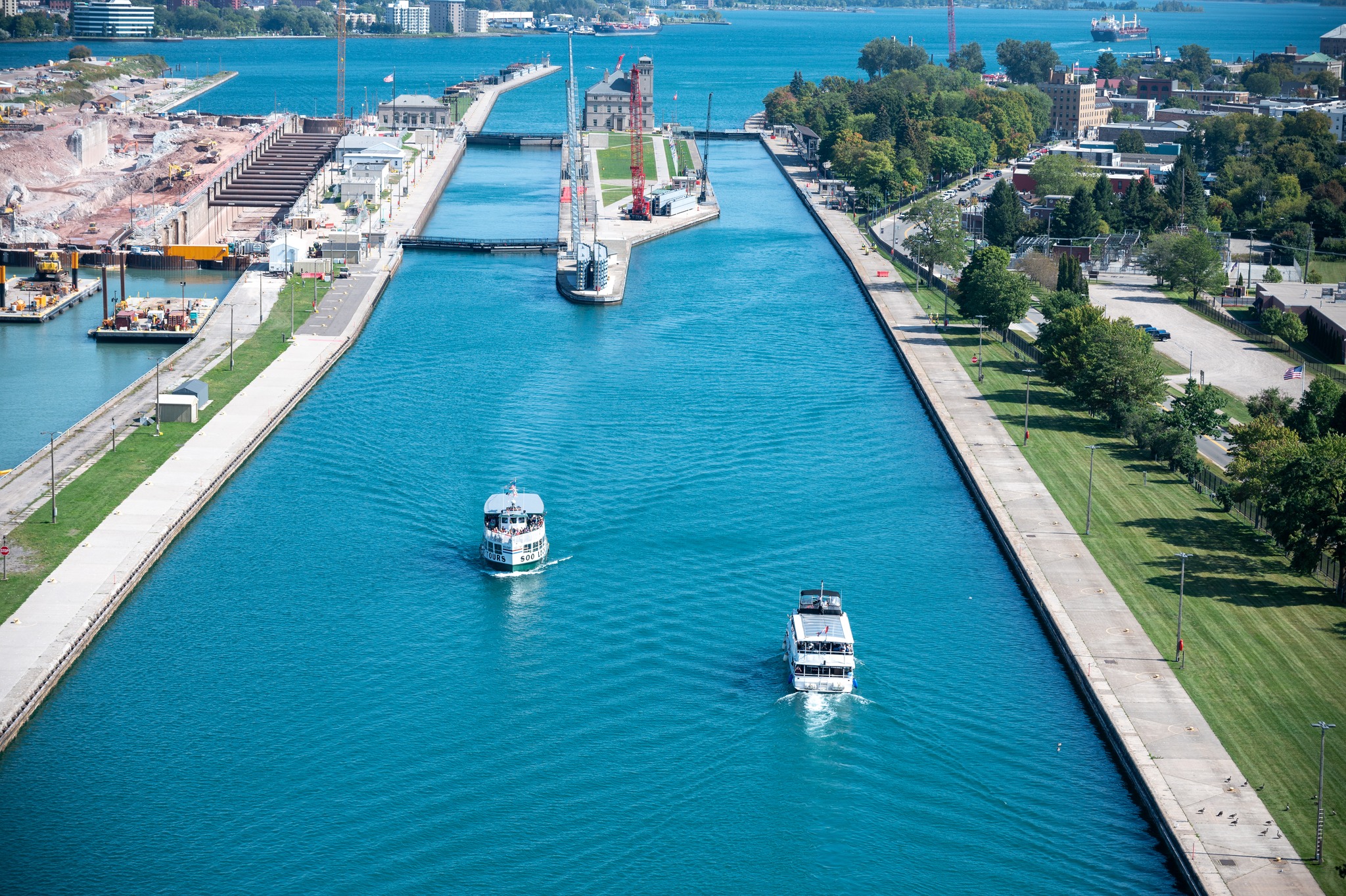  passenger cruise ships sail calmly through the locks in the azure water of Sault Ste. Marie, green trees and grass surround the vibrant waterfront boardwalk. A bustling downtown sits on the skyline.