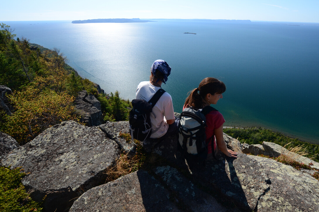 two people sit a the top of a rock cliff overlooking the broad shining blue expanse of Lake Superior, with Sleeping Giant on the horizon. The trees along the cliff face are turning yellow for autumn.