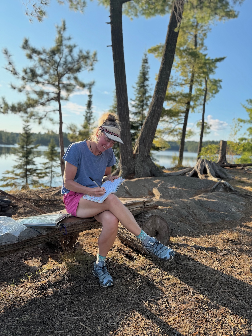 Woman sitting on a bench with lake and trees in background, writing in journal.