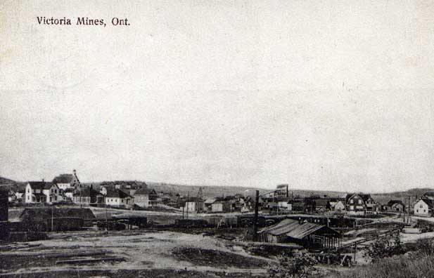a historic black and white photograph of the town of Victoria Mines, a tiny village of a few wooden houses surrounded by shrubs and rocky terrain.