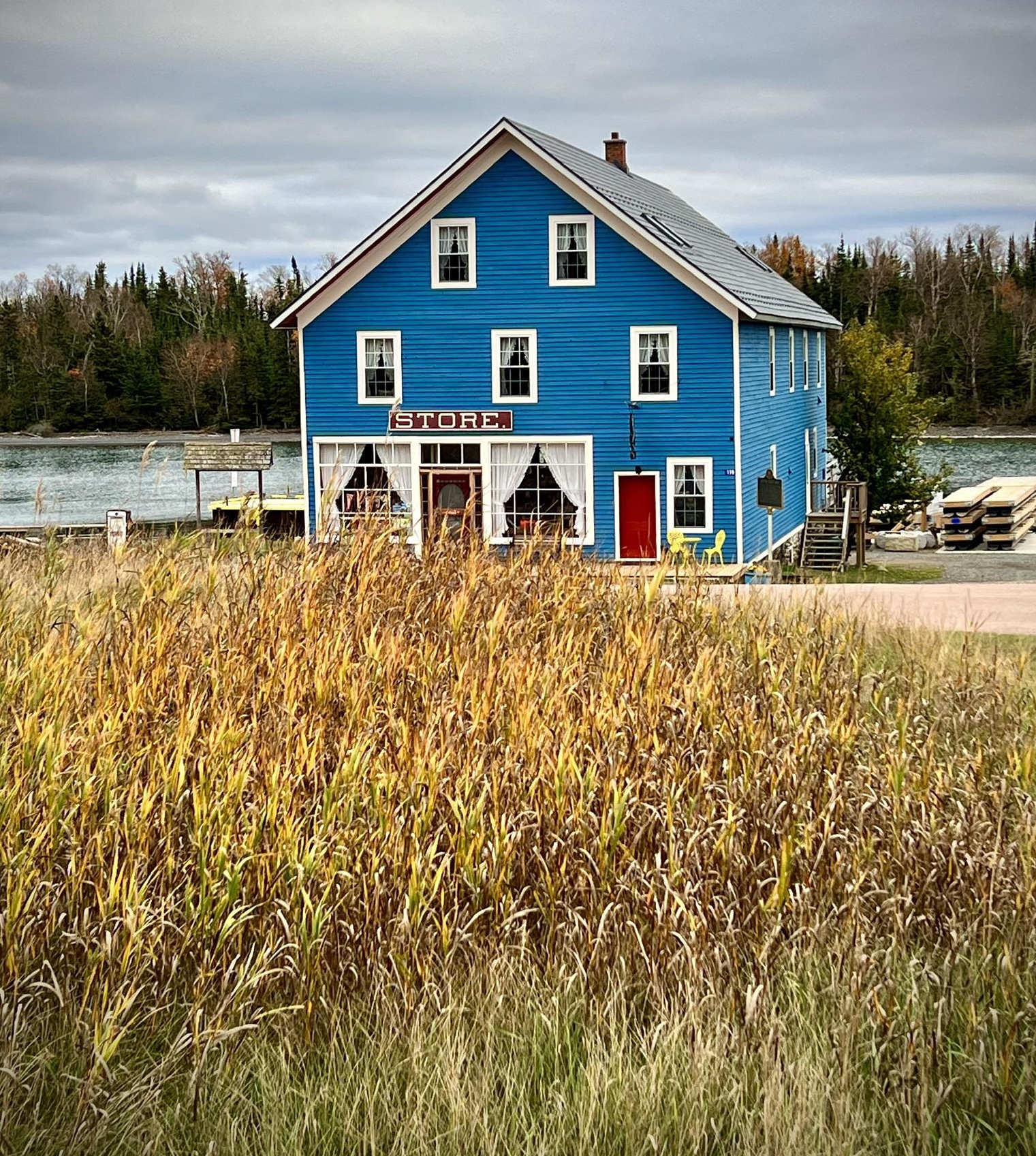 The Silver Islet General Store; a large blue antique building with wooden porch next to a bay, with broze autumn grass waving in the field in front under a grey autumn sky. 