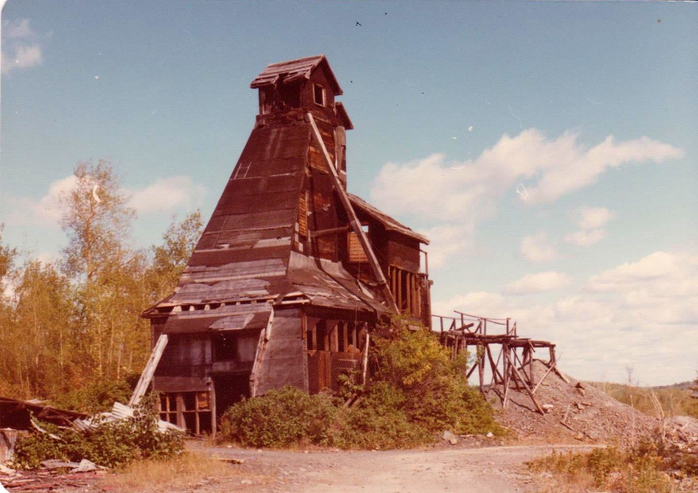 An old colourized photo of a delapidated wooden mine head surrounded by brown brush and a pale sky.