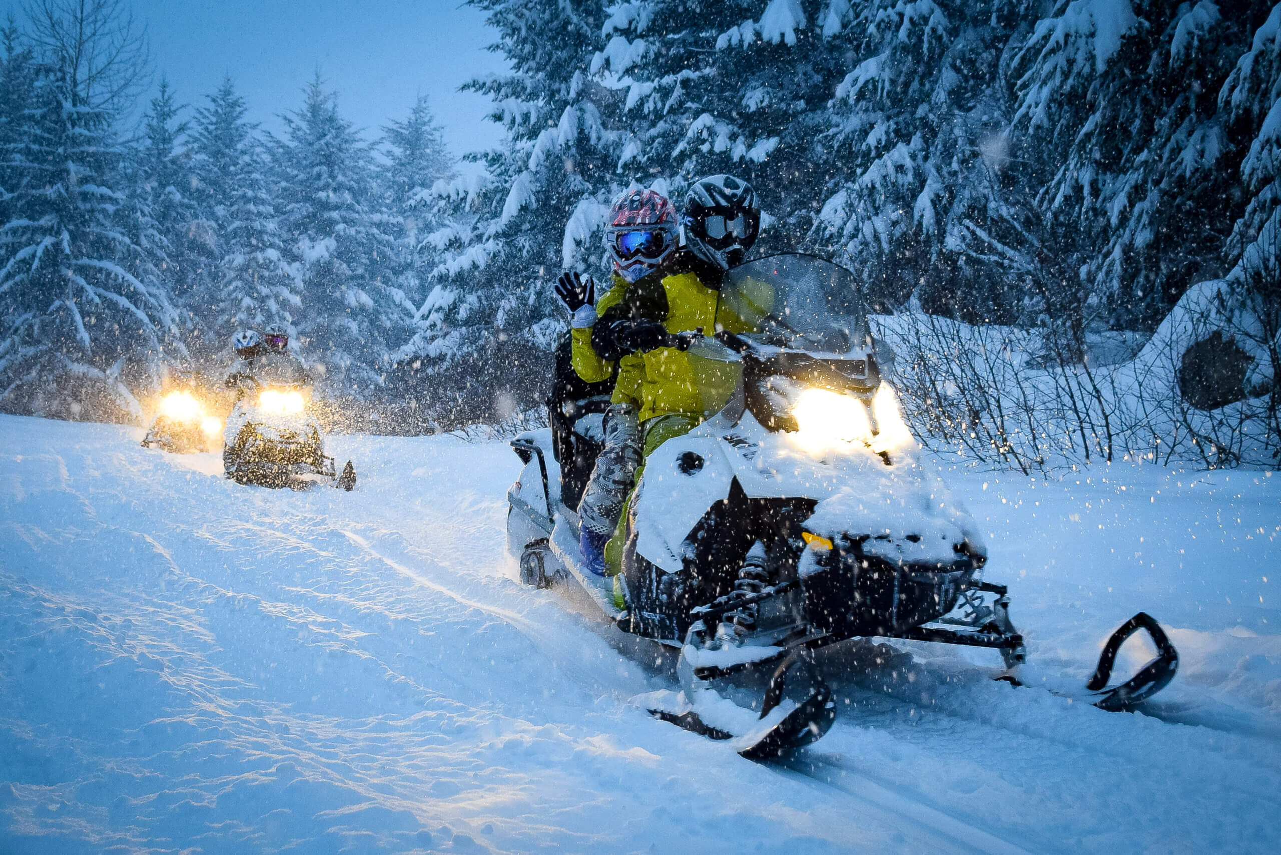 a group of people on snowmobiles ride through a snowy forest at twilight, with the trail illuminated by their headlights. 