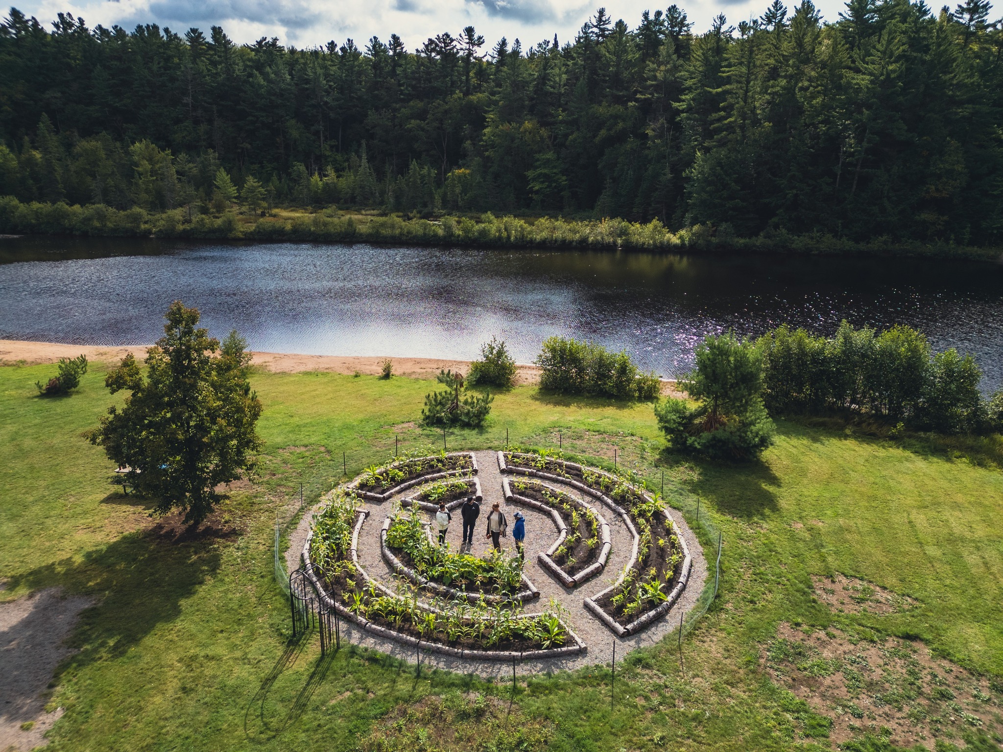 a circular garden with carefully laid beds and gravel paths, surrounded by lush green grass and forest and a river. 