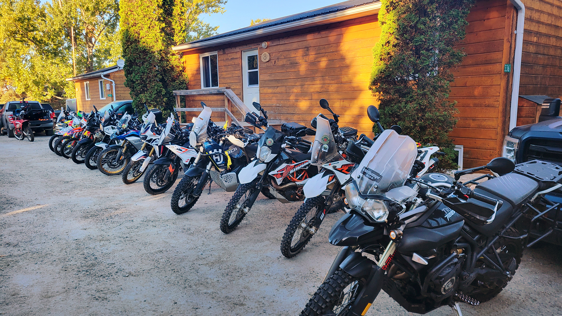 a series of motorcycles lined up and parked together alongside a wooden building on a sunny morrning, with bright fall trees in the background.