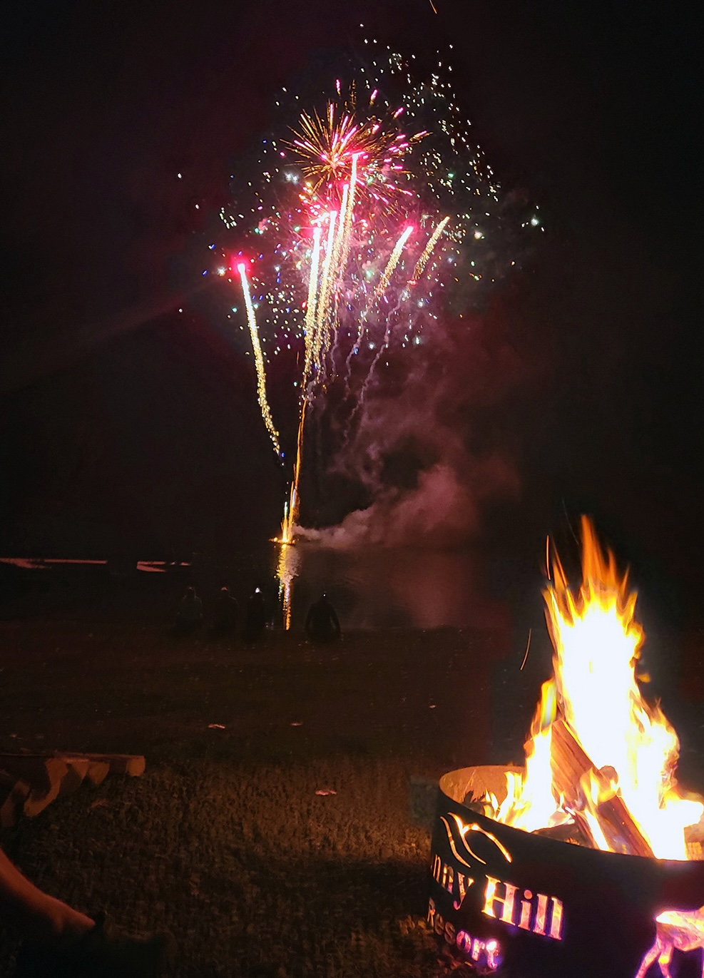 red fireworks shoot into the dark night sky while a campfire in a metal fire ring labelled "Sunny Hill Resort" burns brightly below. 