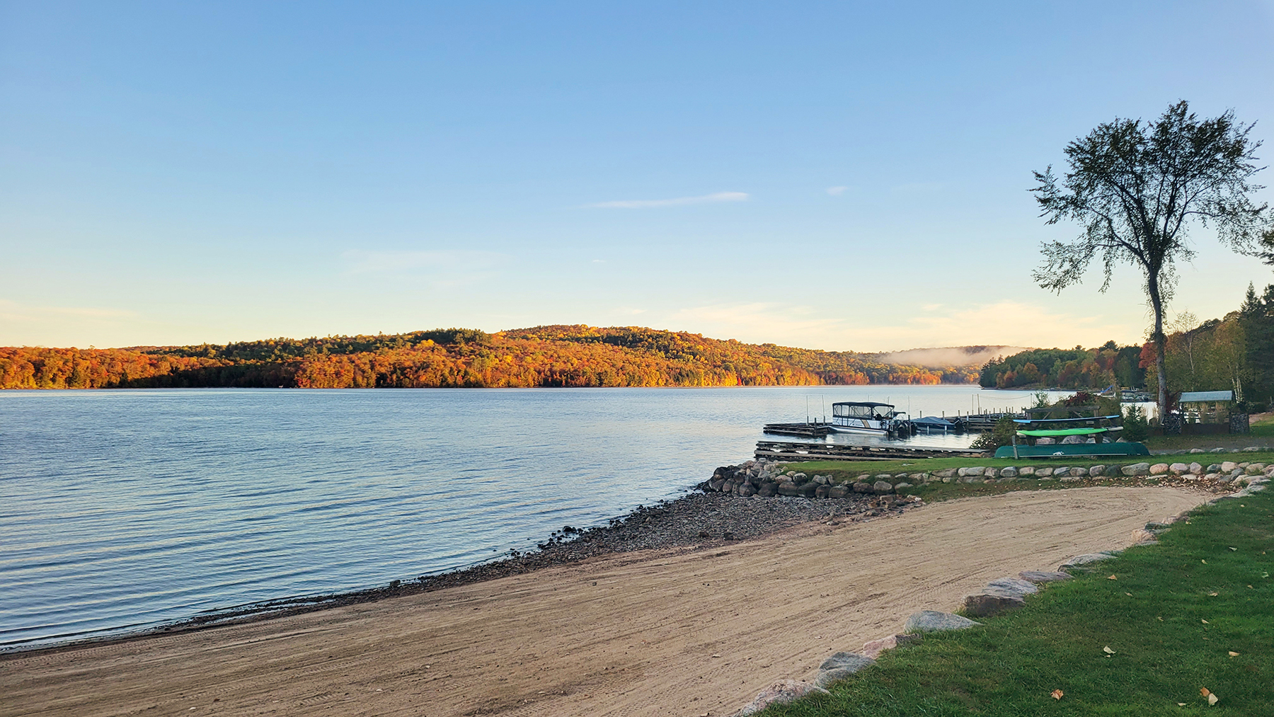 the sun dawns on a perfectly clear morning over the calm blue water of the Madawaska. A soft sandy beach, green trees and boat launch line the bank. 