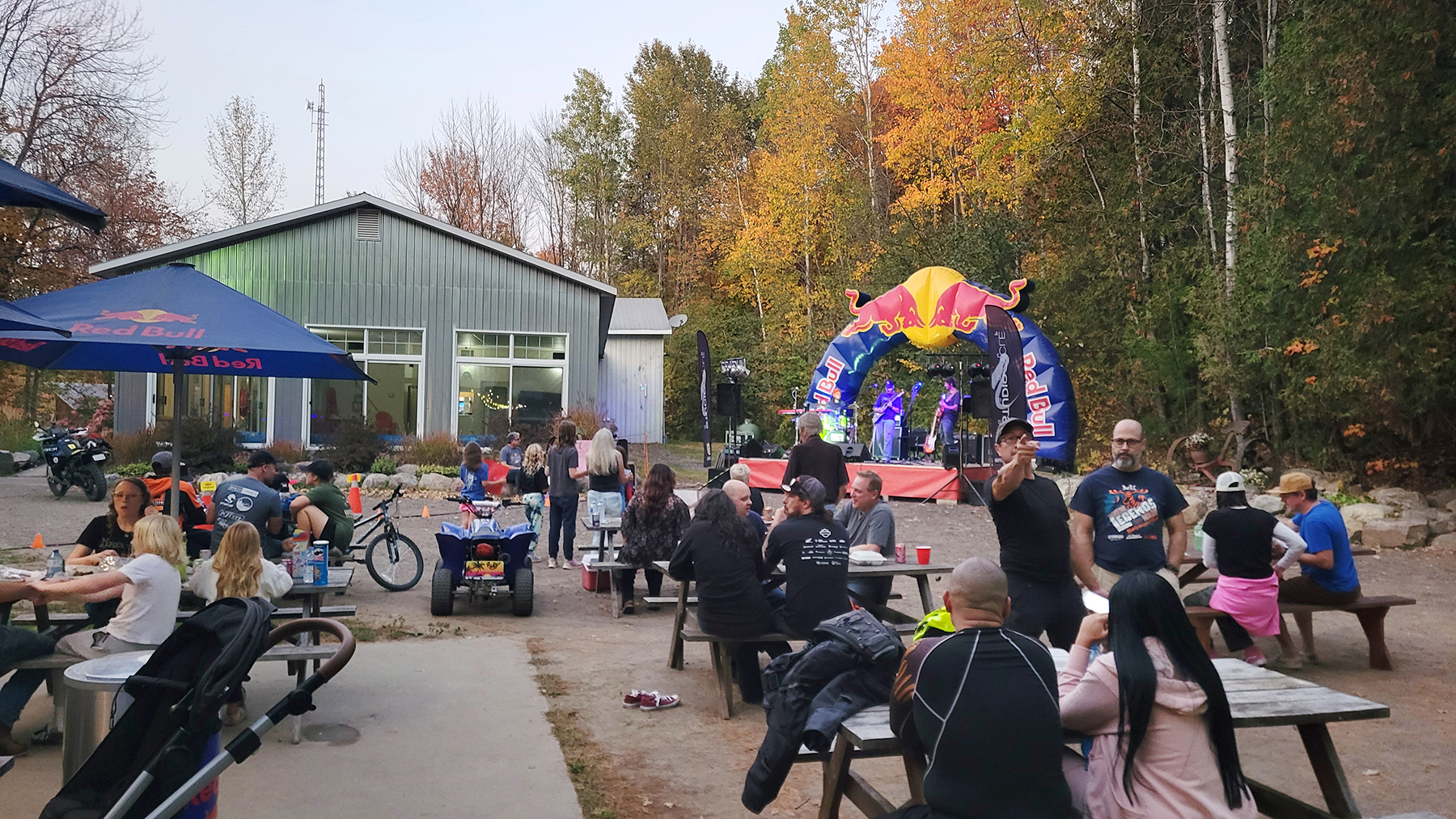 outdoor tables filled with diners and an inflatable arch over a band stage at one end. Colourful autumn forest surrounds the dining area. 