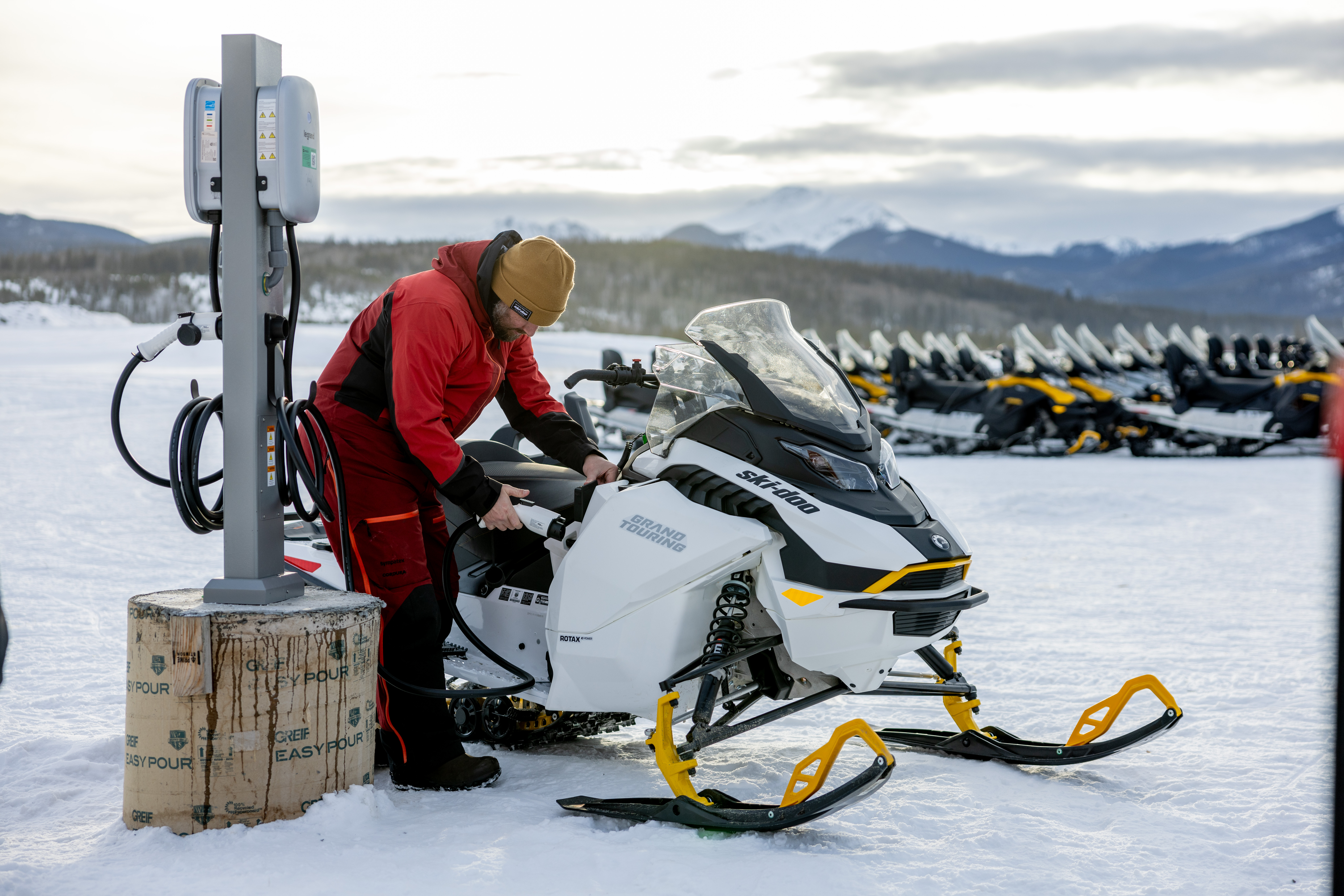 a person in winter gear plugging in their electric snowmobile at a charge station on a winter day. 