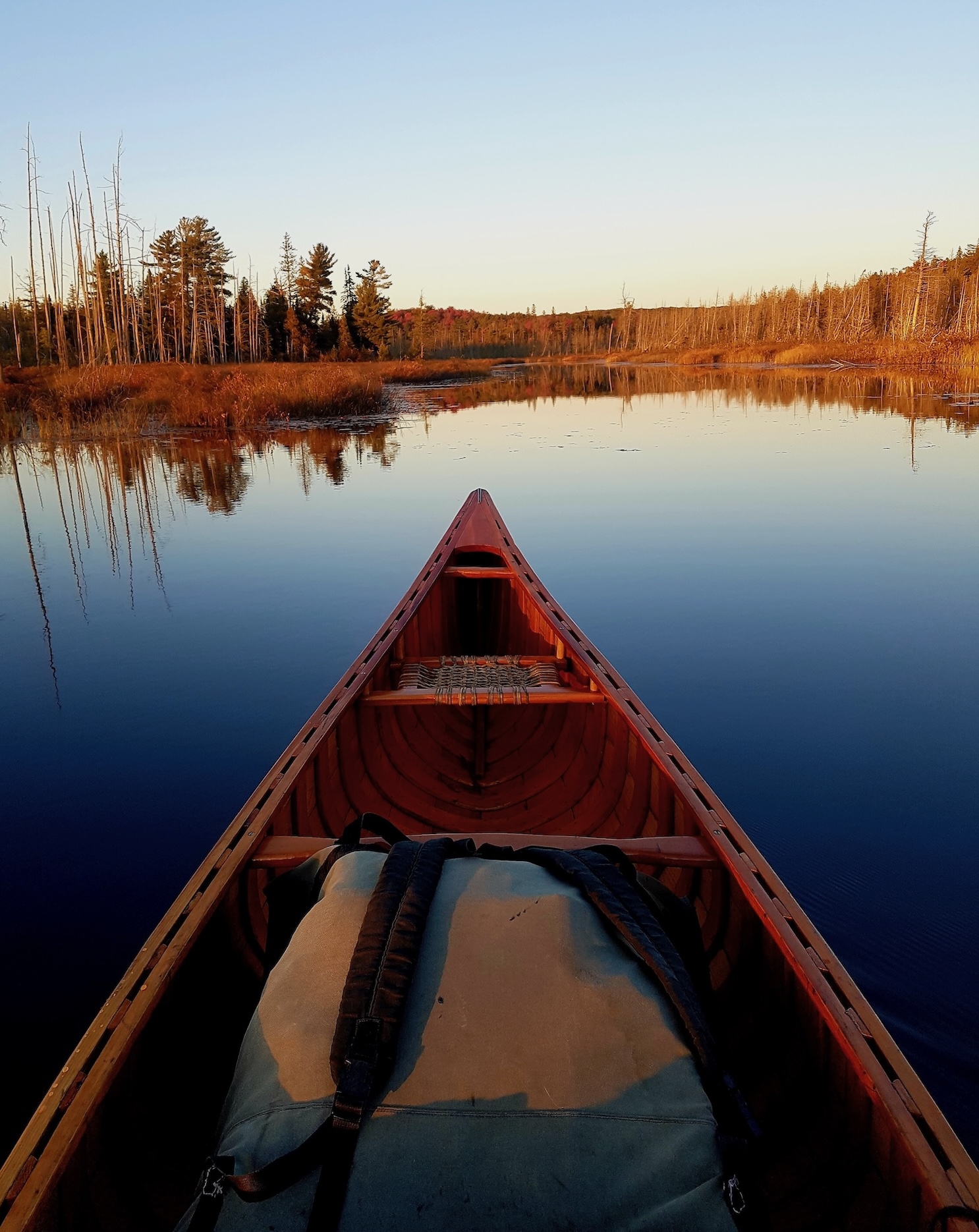a canoe with a canvas pack at the bow pointing out into a glassy blue lake reflecting a clear blue autumn sky. 