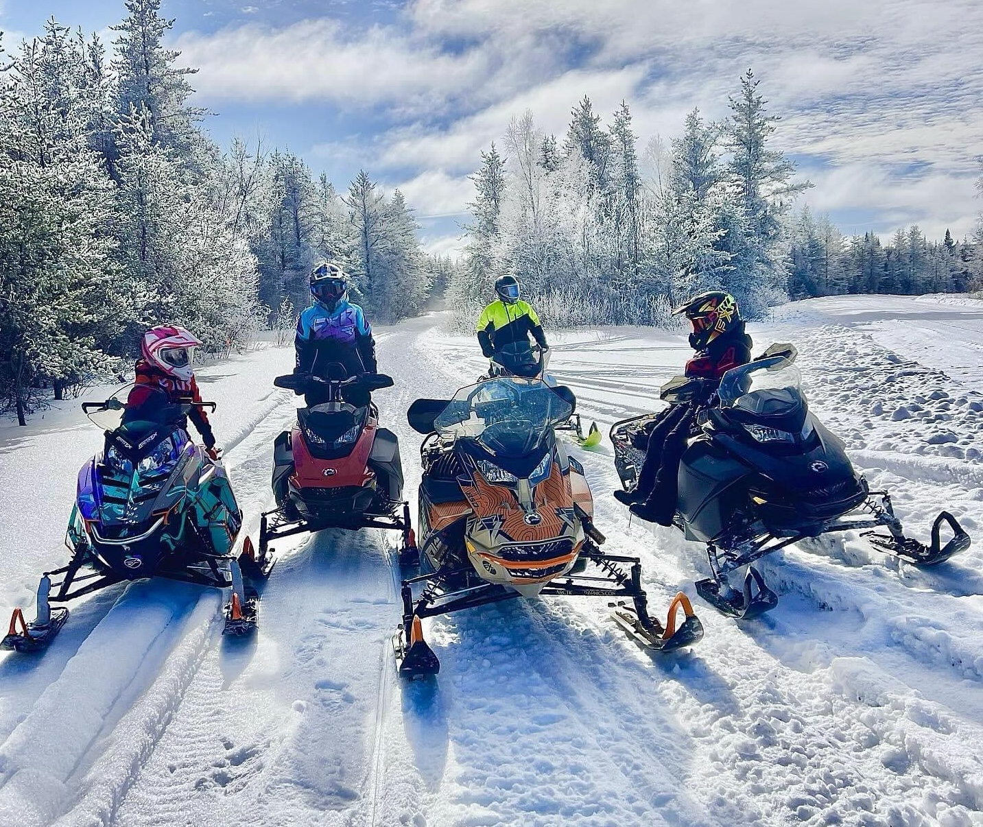 4 snowmobilers parked on the trail side by side, glistening powder shining in the sun on the forest all around. 