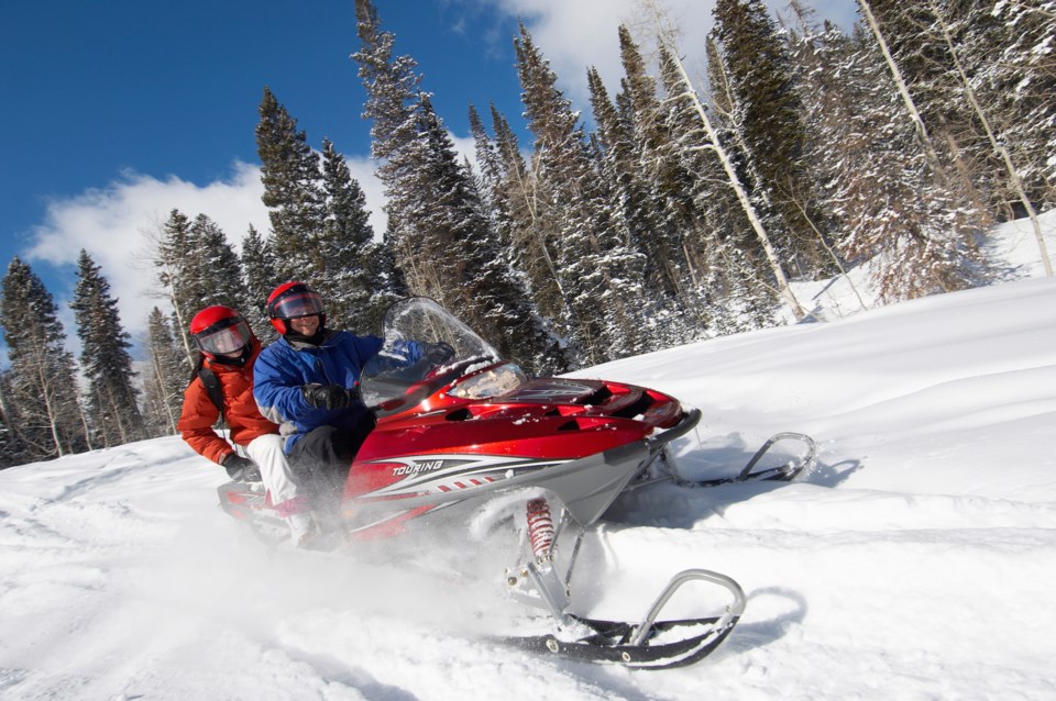 two young people on a snowmobile cruise along a snowy trail on a sunny winter day, kicking up powder with their ski. Evergreen forest is in the background. 