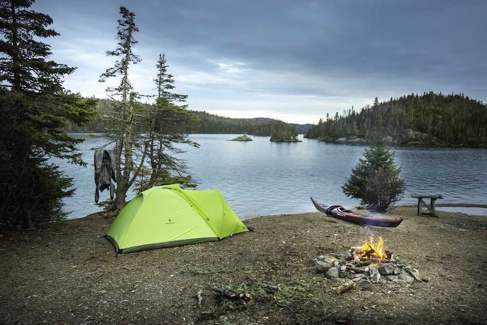 Green tent with sea kayak pulled up on shore next to it. Campfire in foreground. Lake in background.