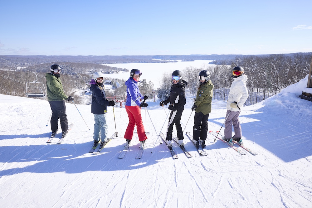 Six people on skis at top of hill with frozen lake in background