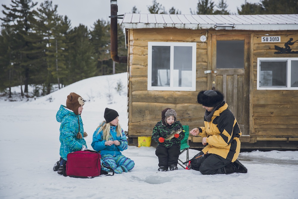 Family sitting outside fishing hut fishing in a hole in the ice