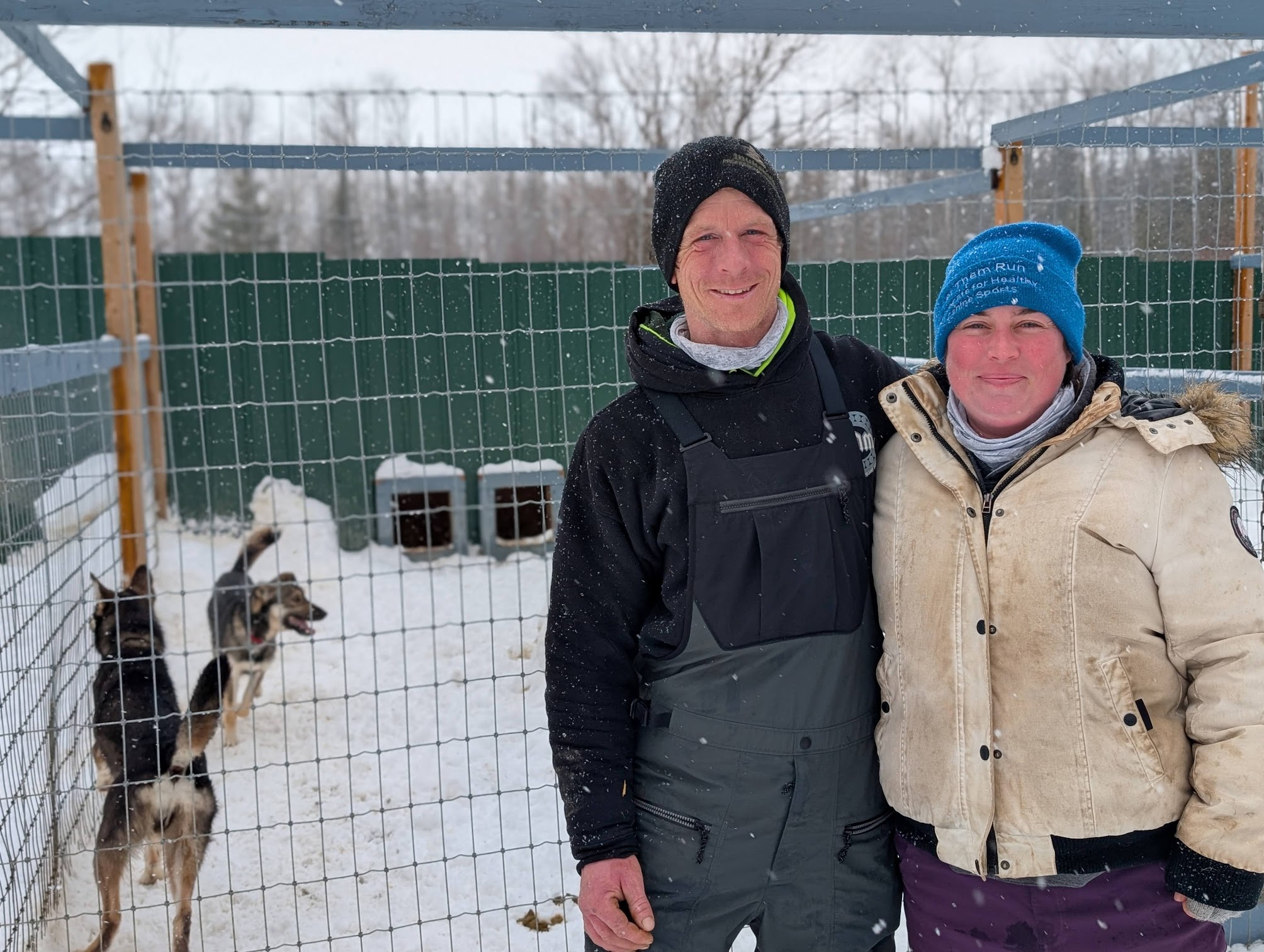 Abitibi Sled Dog owners Jacob and Chantale smile with arms around each other's shoulders, standing in front of their team of huskies in a large snowy kennel. 