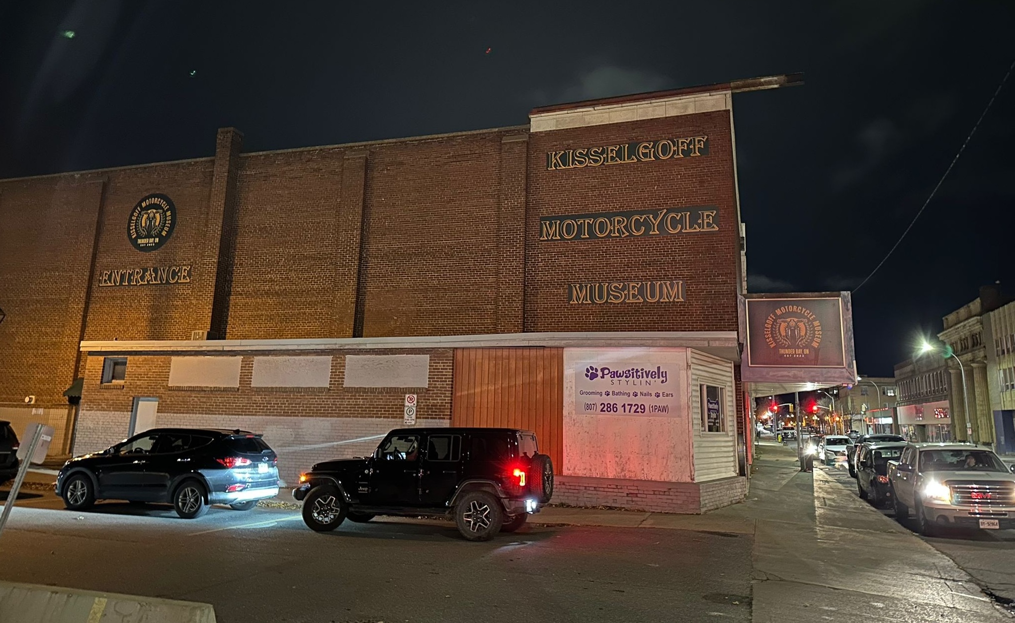 The Kisselgoff Motorcycle Museum in Thunder Bay lit up at night and surrounded by cars parking outside. The museum is housed in a tall 1948 brick building that once served as a theatre.