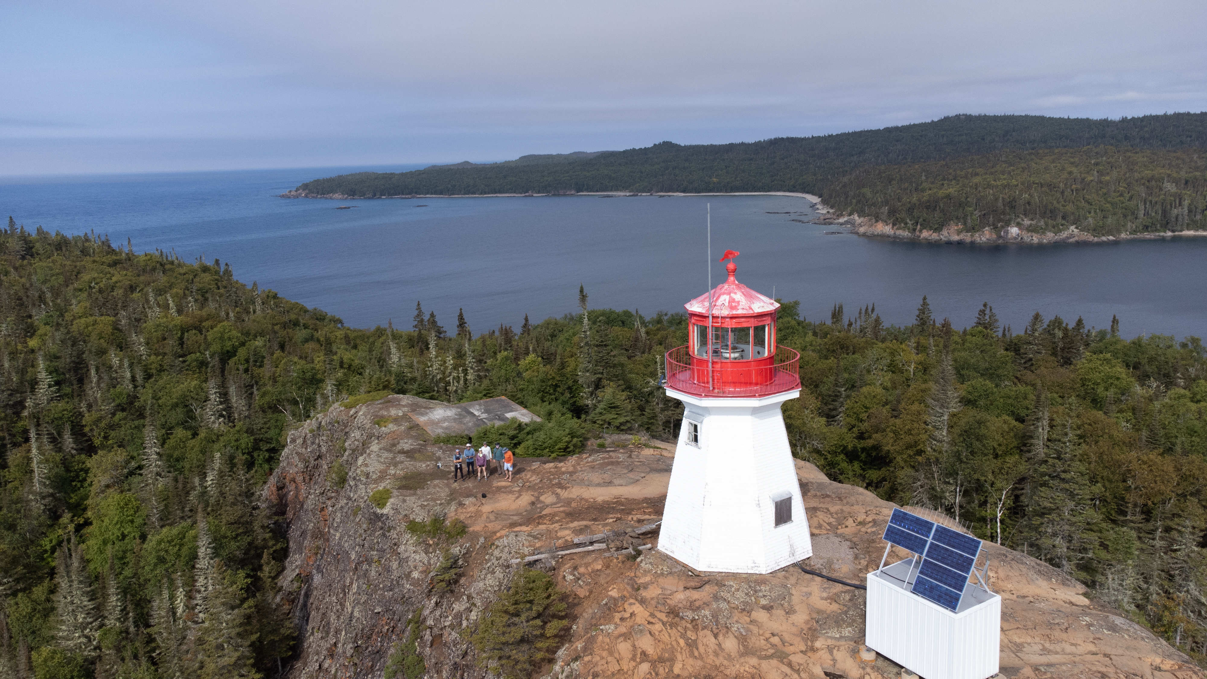 Drone shot of people standing at edge of cliff beside red and white lighthouse