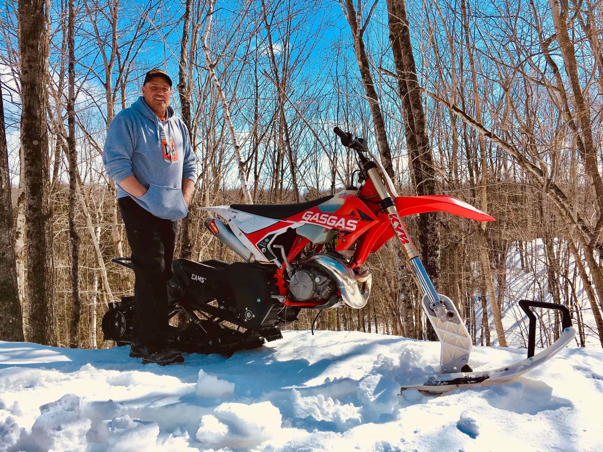 Garage owner Mark Williams smiling next to a converted tracked motocross snowbike on a snowy forest trail on an sunny winter day. 