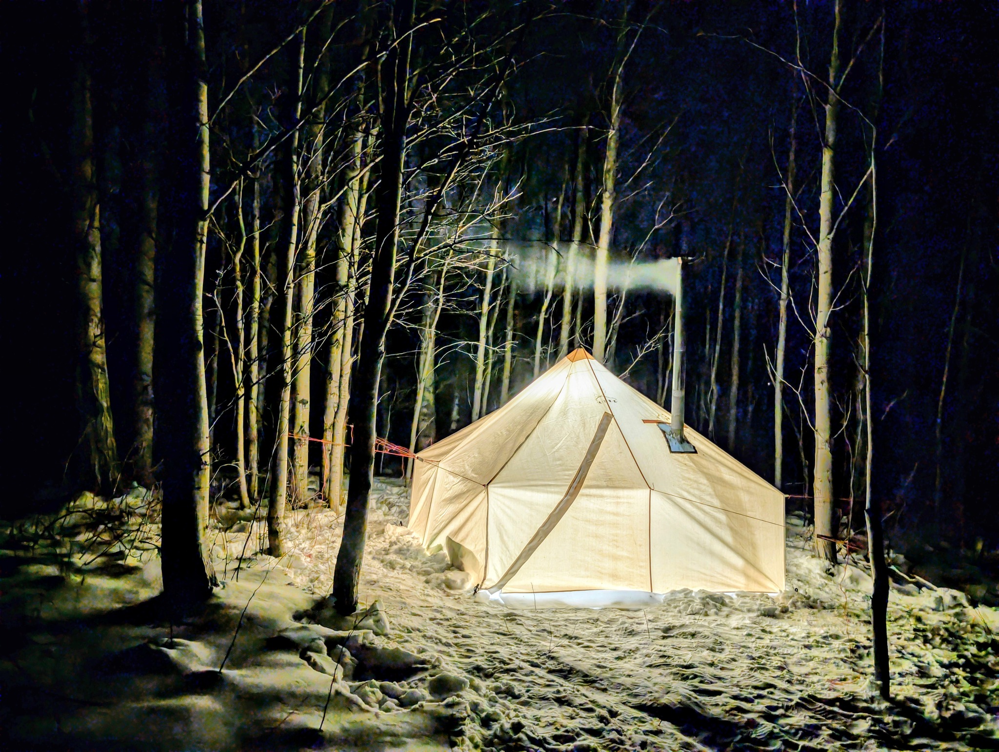 a canvas prospector's tent glows like a lantern in the night, illuminated from within, in a dark snowy forest. Smoke wisps out of its metal chimney.