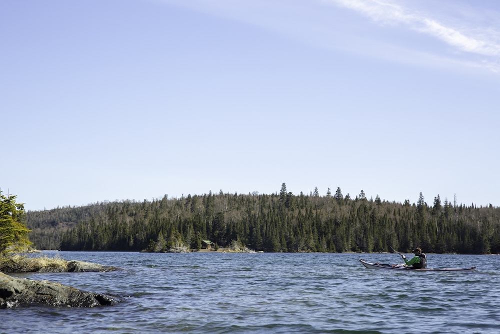 Man in sea kayak with small cabin on distant shore in background