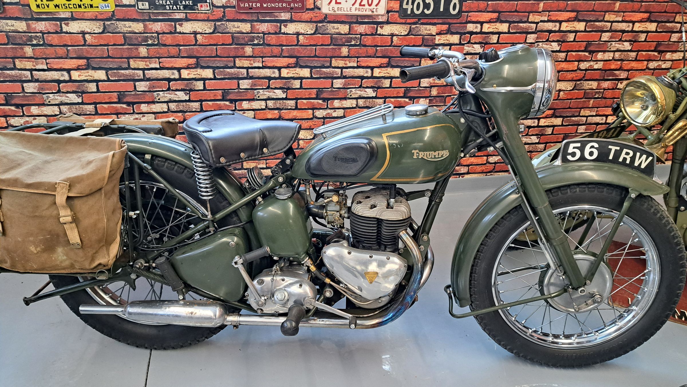 A vintage olive green Triumph military motorcyle with leather saddlebags in front of a brick wall at the Kisselgoff Motorcycle Museum.
