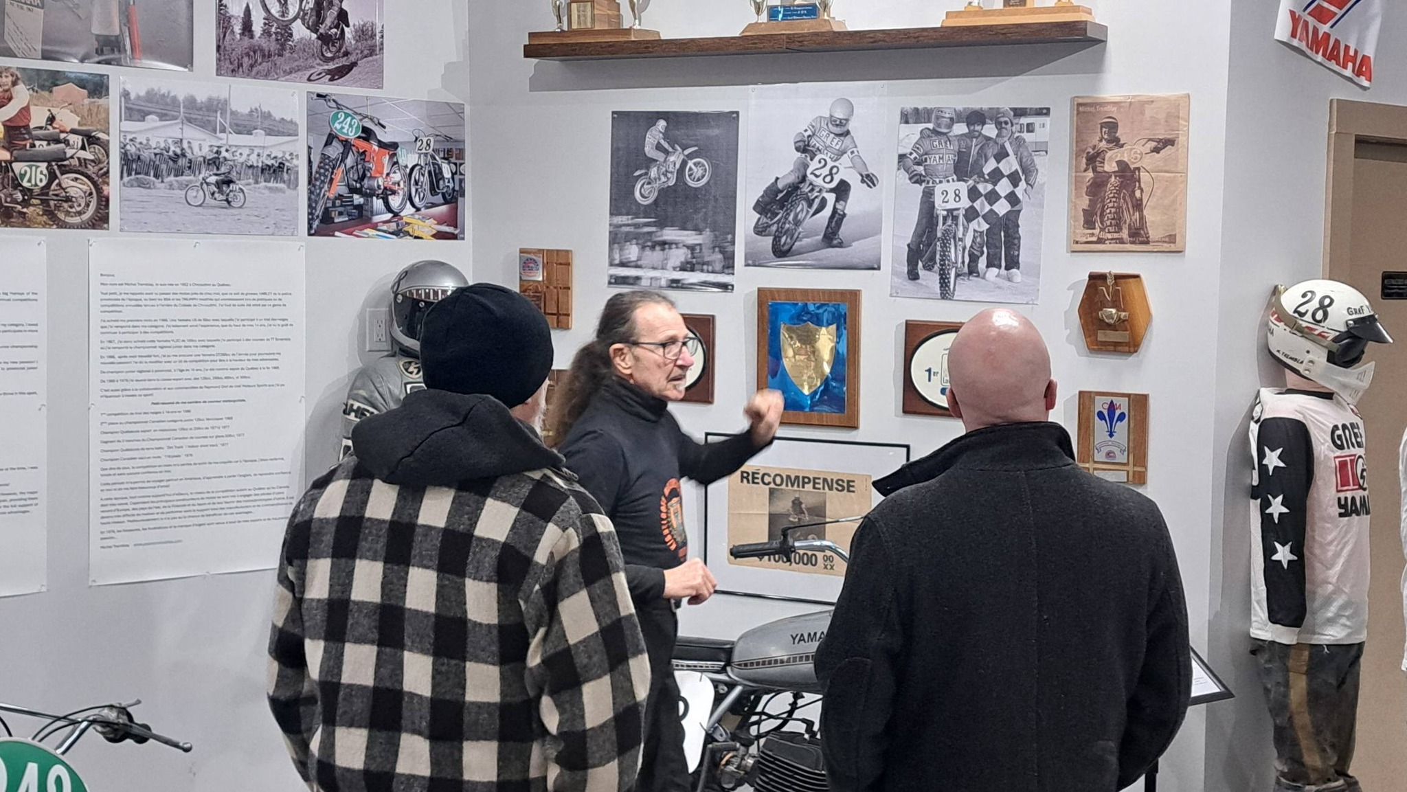 a staff member speaks to to curious visitors about the motorcycle memorabilia and photos on a wall at the Kisselgoff Motorcycle Museum.
