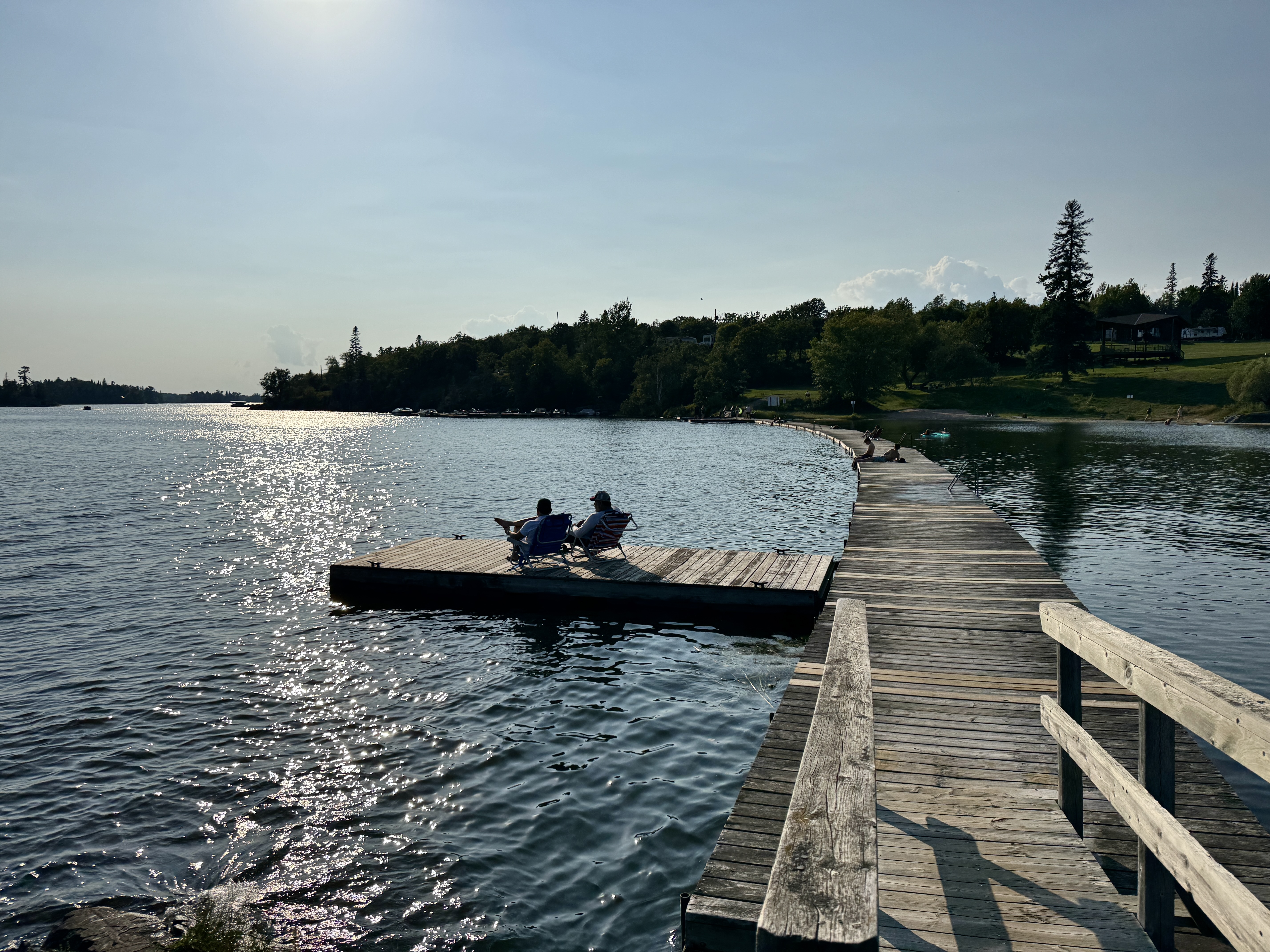 two people sit on a long wooden pier surrounded by a deep lake on a sunny afternoon at Anicinabe Park. 
