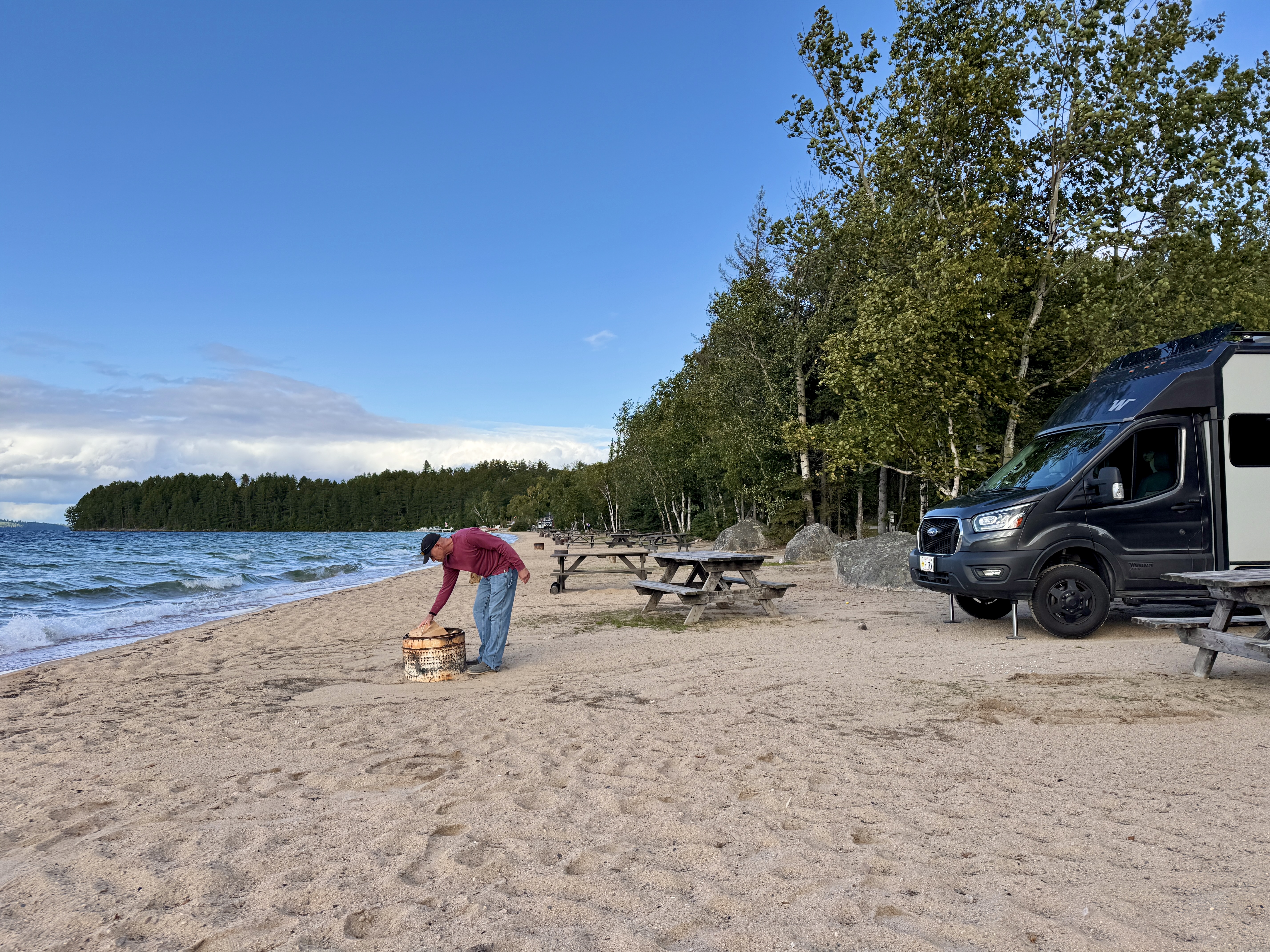 a man picks up firewood off a soft sandy beach surrounded by blue lake and green forest at Browns Clearwater Lodge in Sunset Country. His RV is at the edge of the sand.