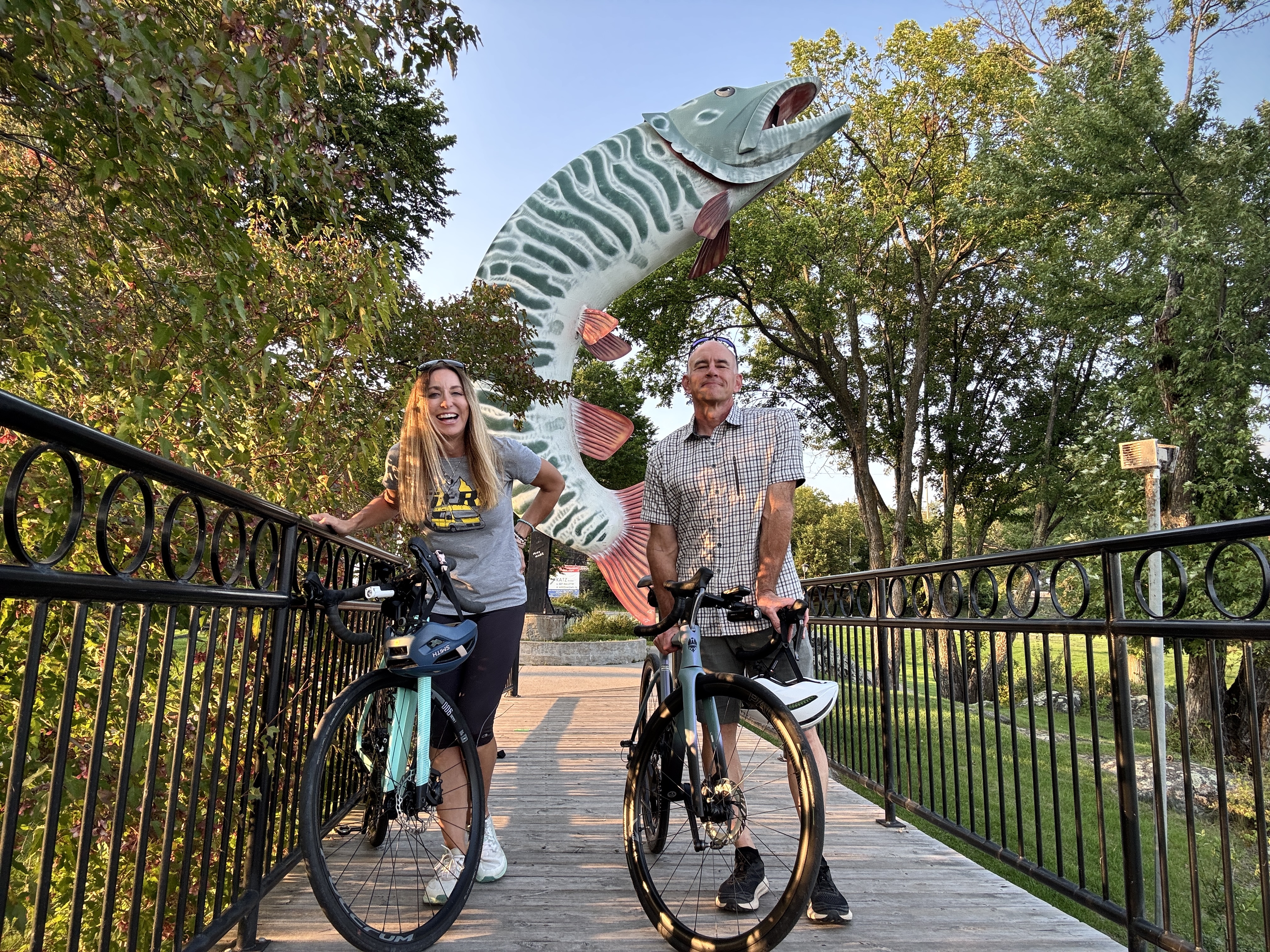 a man and woman on bikes smile in front of a large coloured statue of a muskie in the centre of a forested park in Kenora, Ontario.