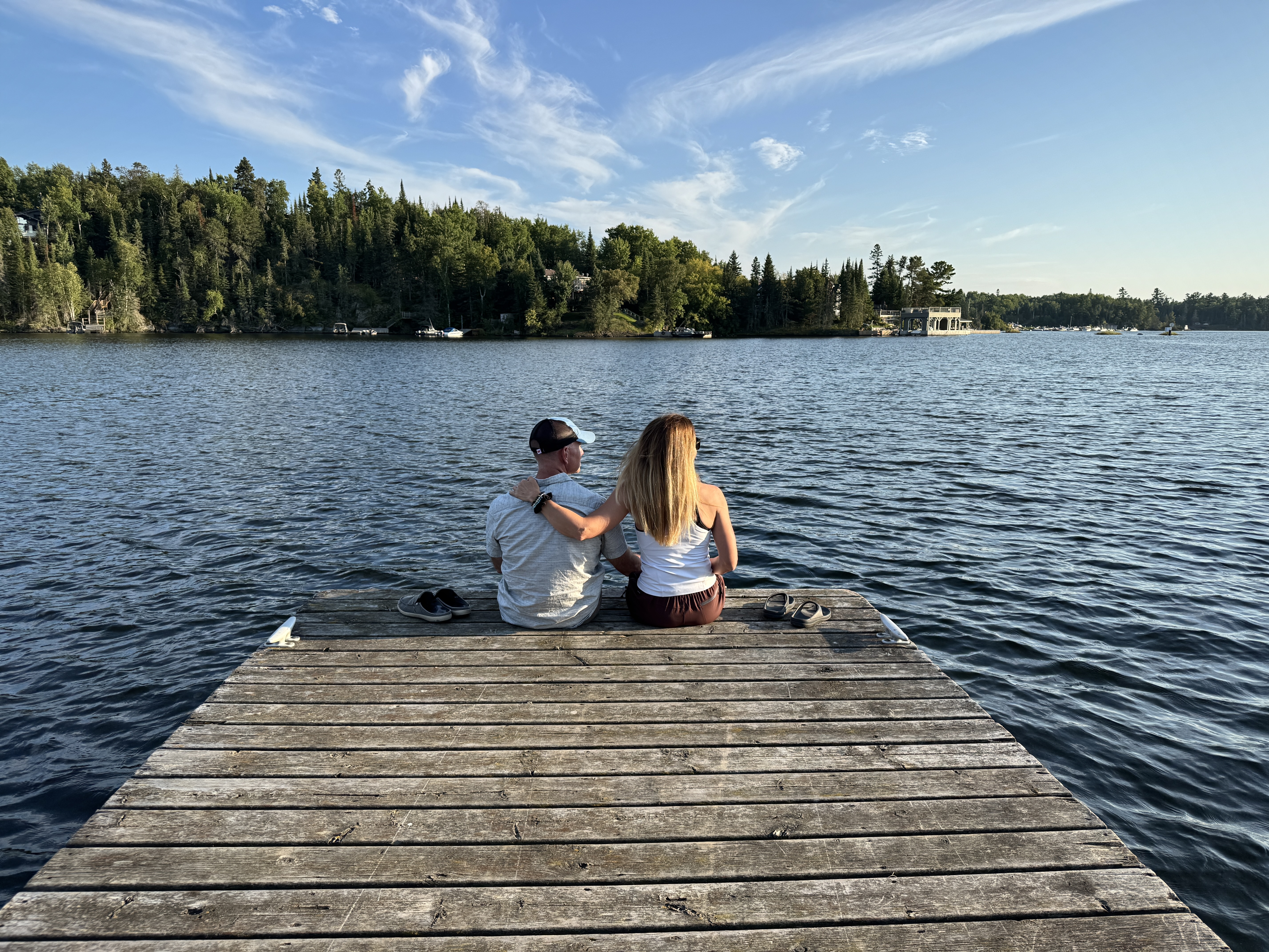 a man and woman sit close together at the end of a wooden dock, looking out across a wide blue lake skirted with green forest under a blue sky. 