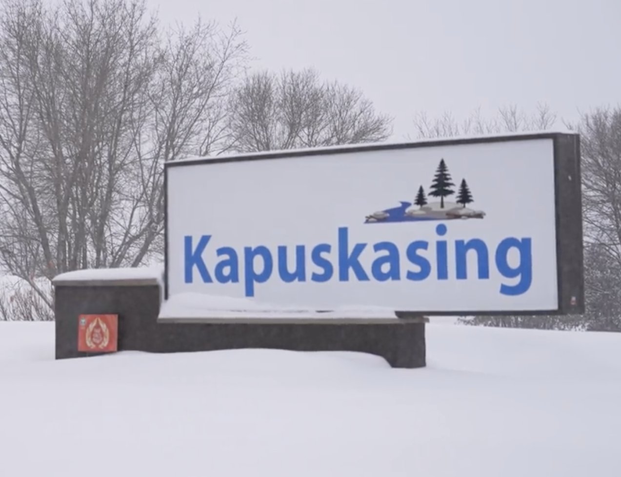 the Kapuskasing town sign, surrounded by deep powdery snow with bare trees and grey sky in the background.