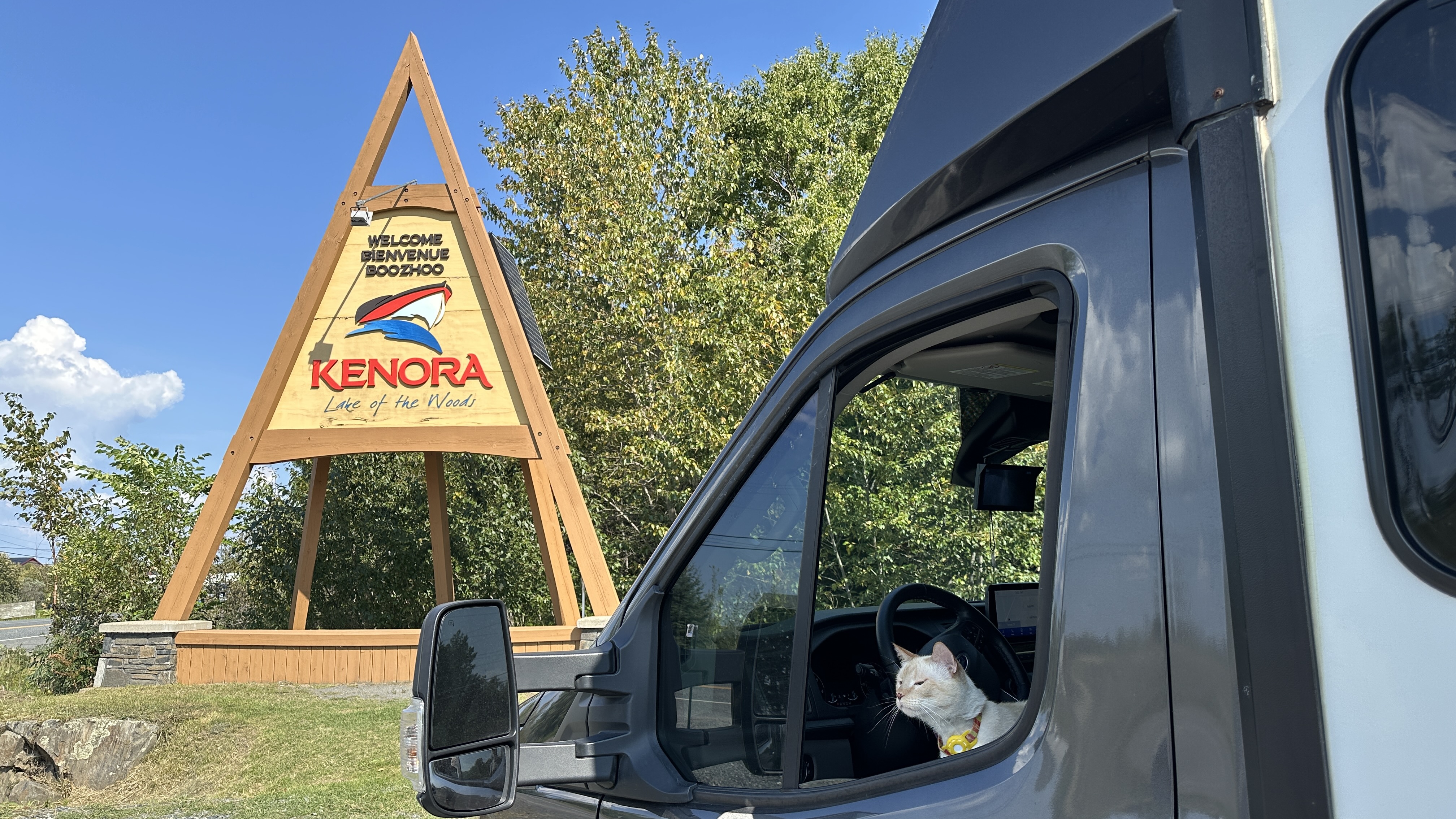 a cat peaks out of the driver's side window of an RV, looking at a large, wooden, pyramid-shaped sign welcoming viistors to Kenora, Northwestern Ontario. Bright blue sky and green trees sit beyond it. 