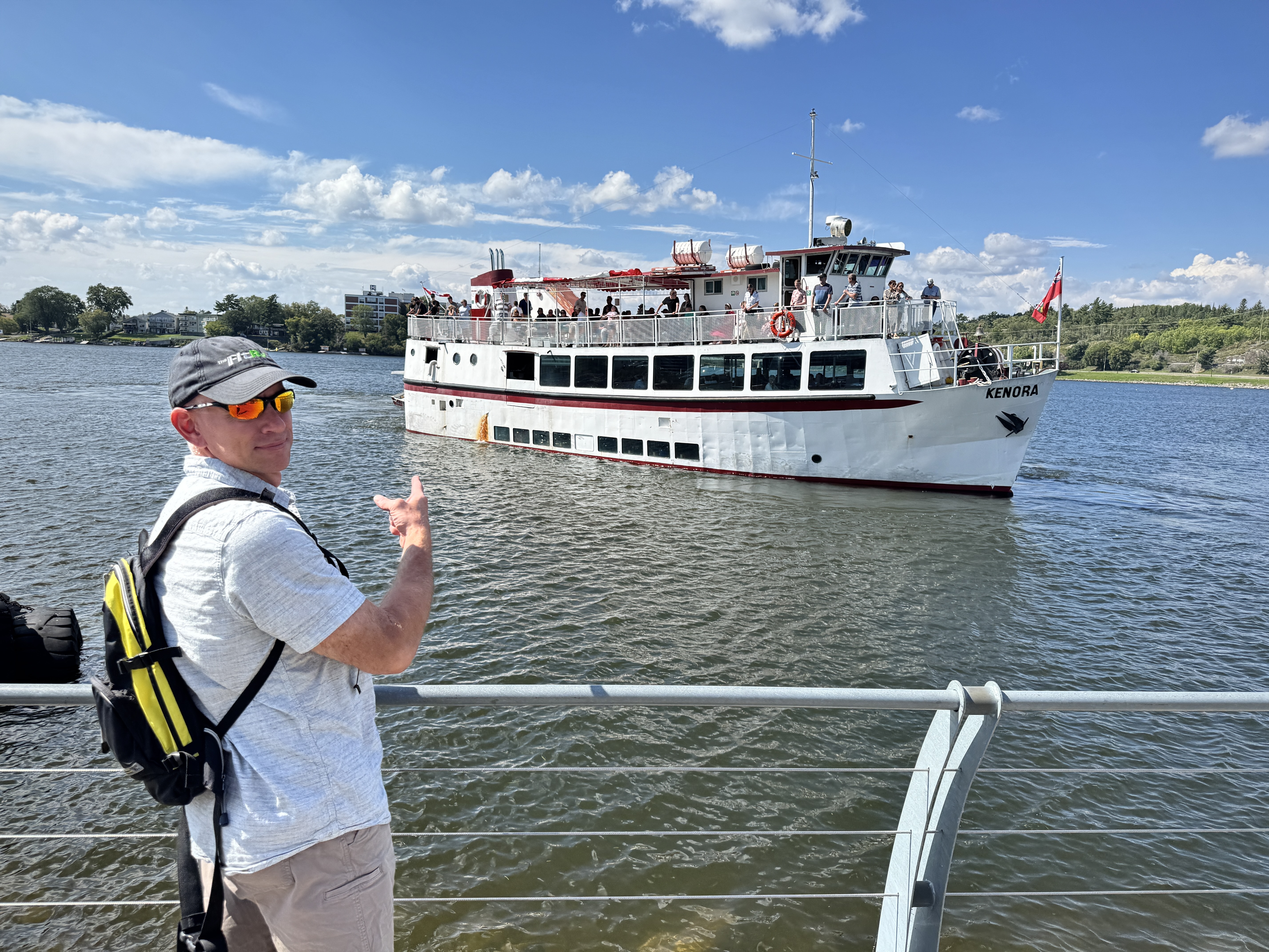 A man wearing a backpack and sunglasses smiles and points at a large lake cruise ship, the MS Kenora, sailing toward him into port on a warm sunny day. 