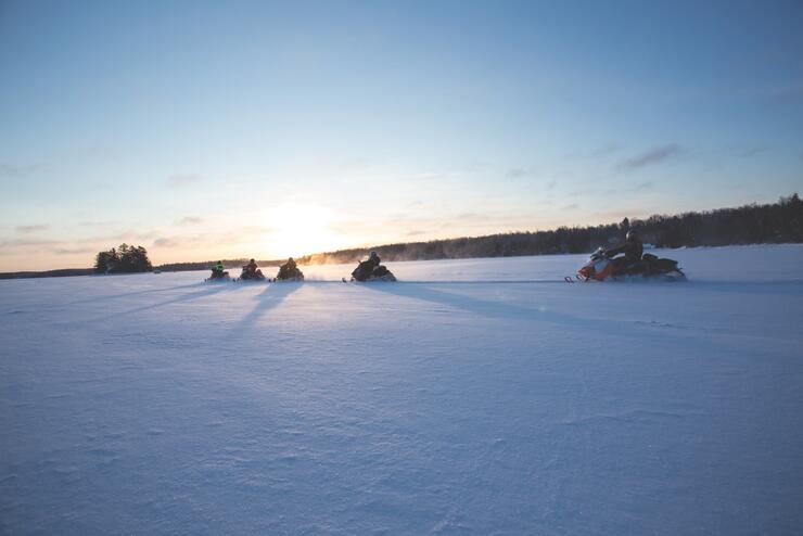 a line of snowmobilers ride across a snowy field in front of a golden setting sun. 