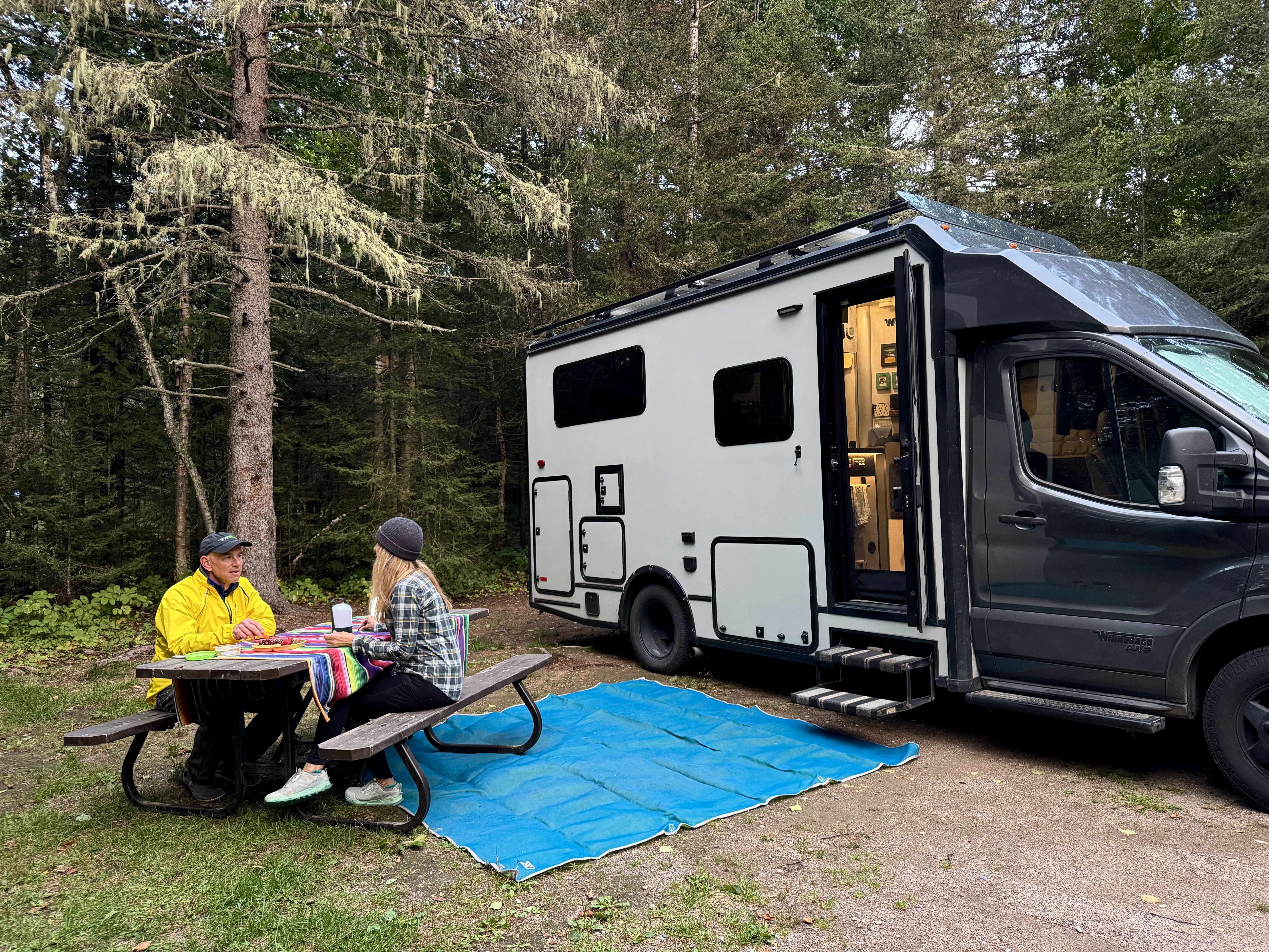 a man and woman sit at a picnic table next to their RV parked at a thickly forested campsite at Whitesand Campground at Rainbow Falls. 