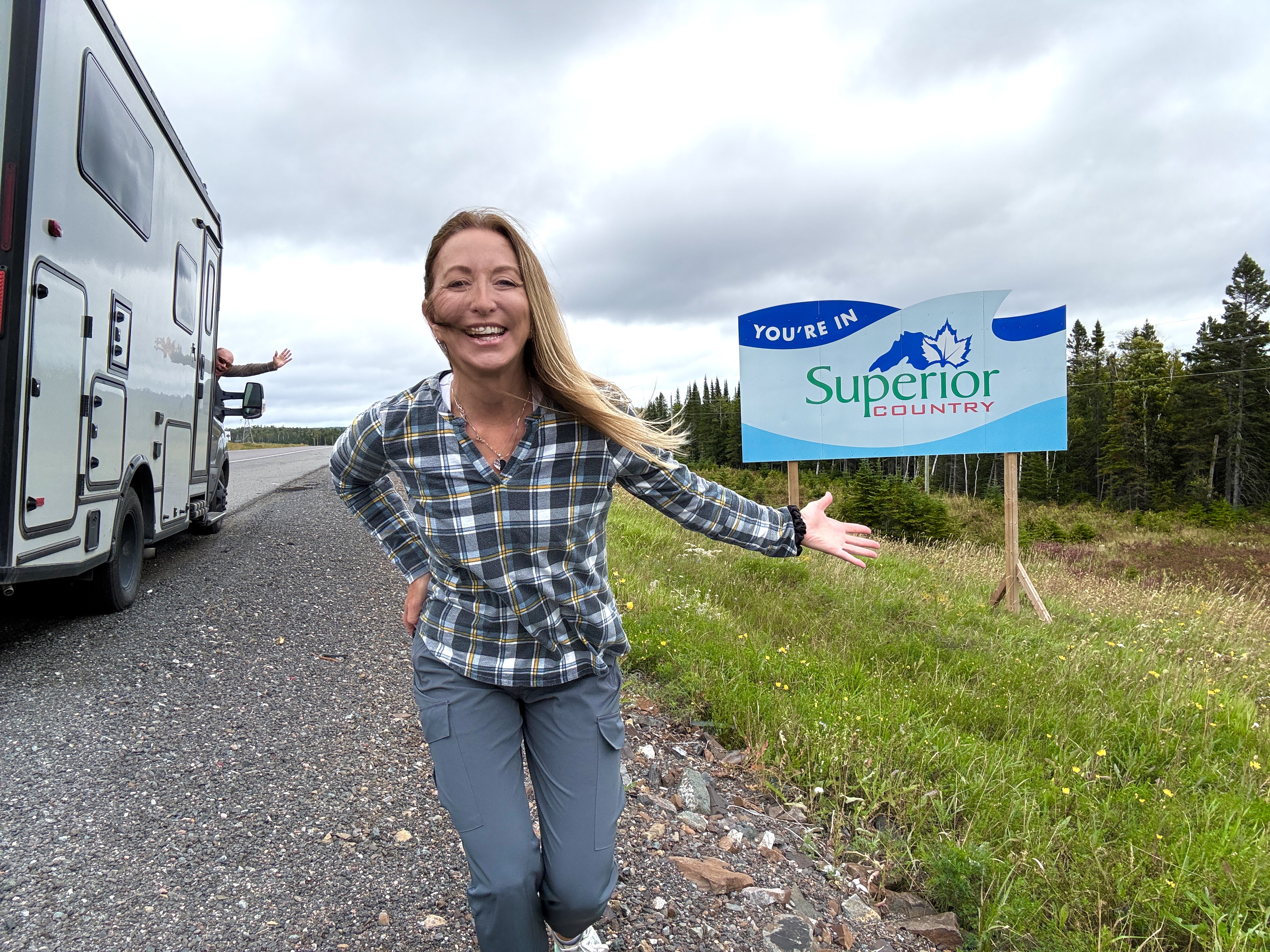 A smiling woman gestures to a roadside sign behind her that reads "You're in Superior Country". Behind her is an RV parked on the side of the road with an arm waving out the window.