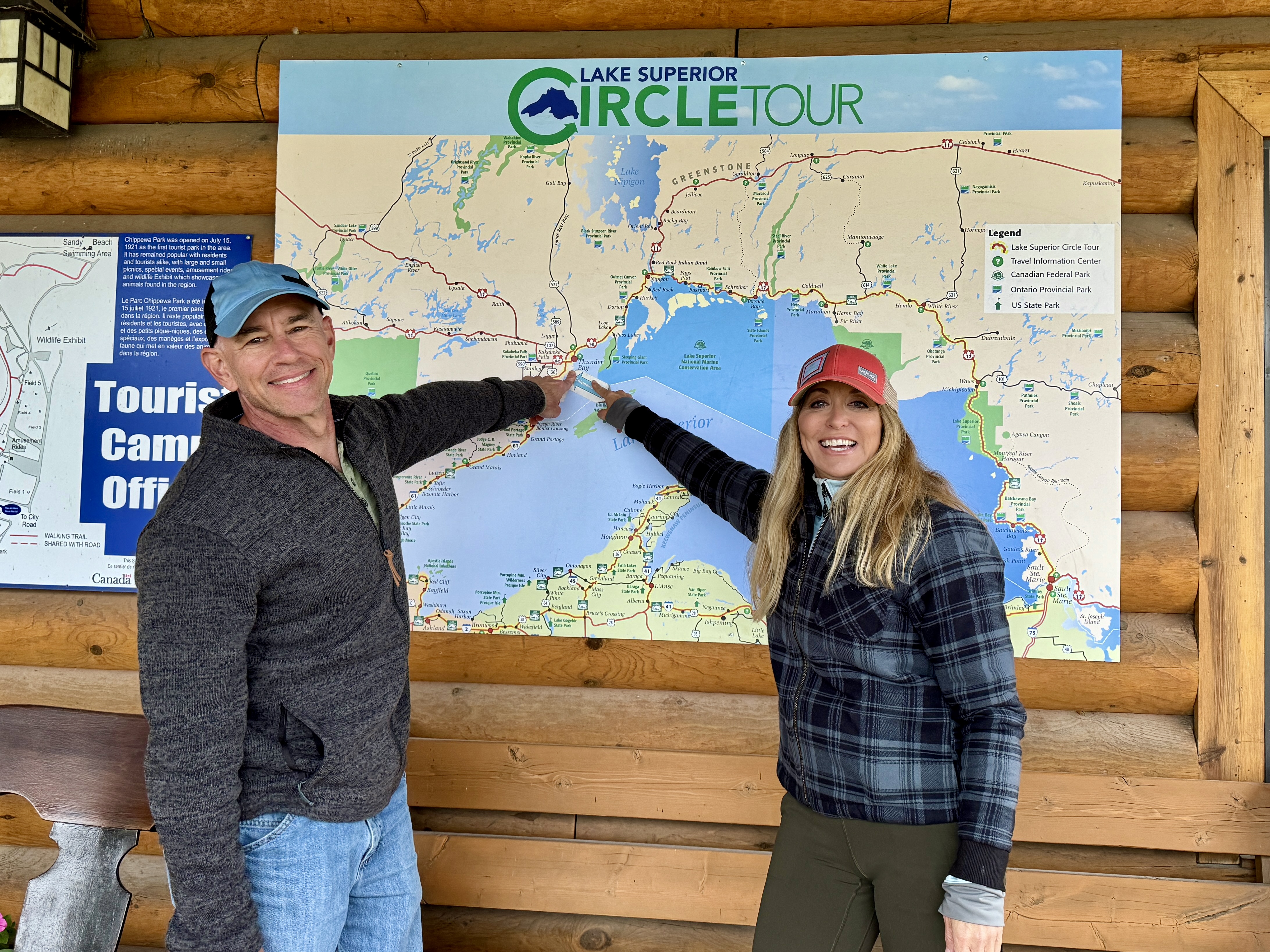 A smiling man and woman stand pointing to a large Lake Superior Circle Tour map on a wooden wall. 
