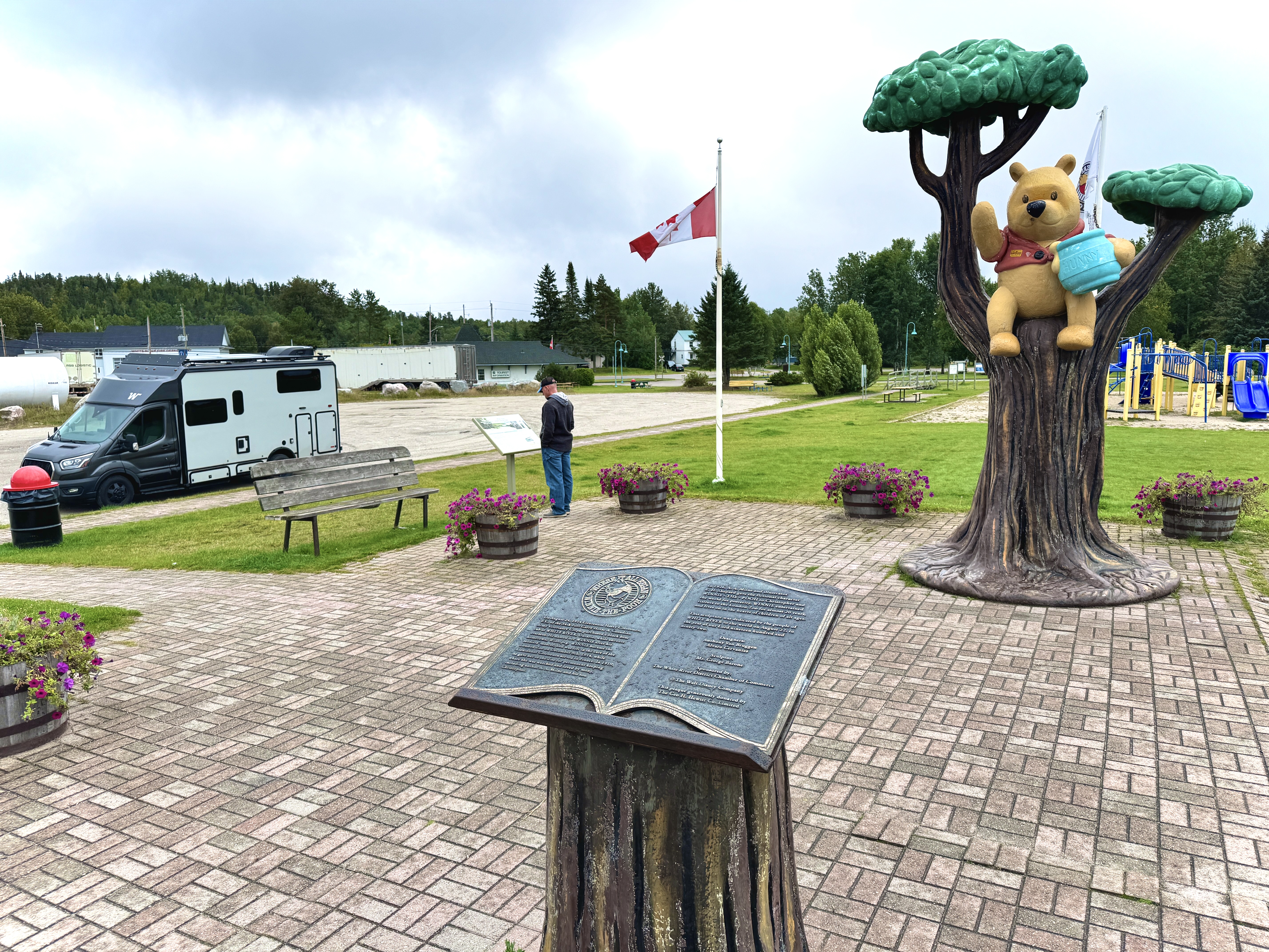 An RV is parked next to the Winnie the Pooh Memorial in White River, Ontario. A man stands on a brick walkway near a large statue of the Disney representation of Winnie the Pooh, holding a honey pot and sitting in a tree. 