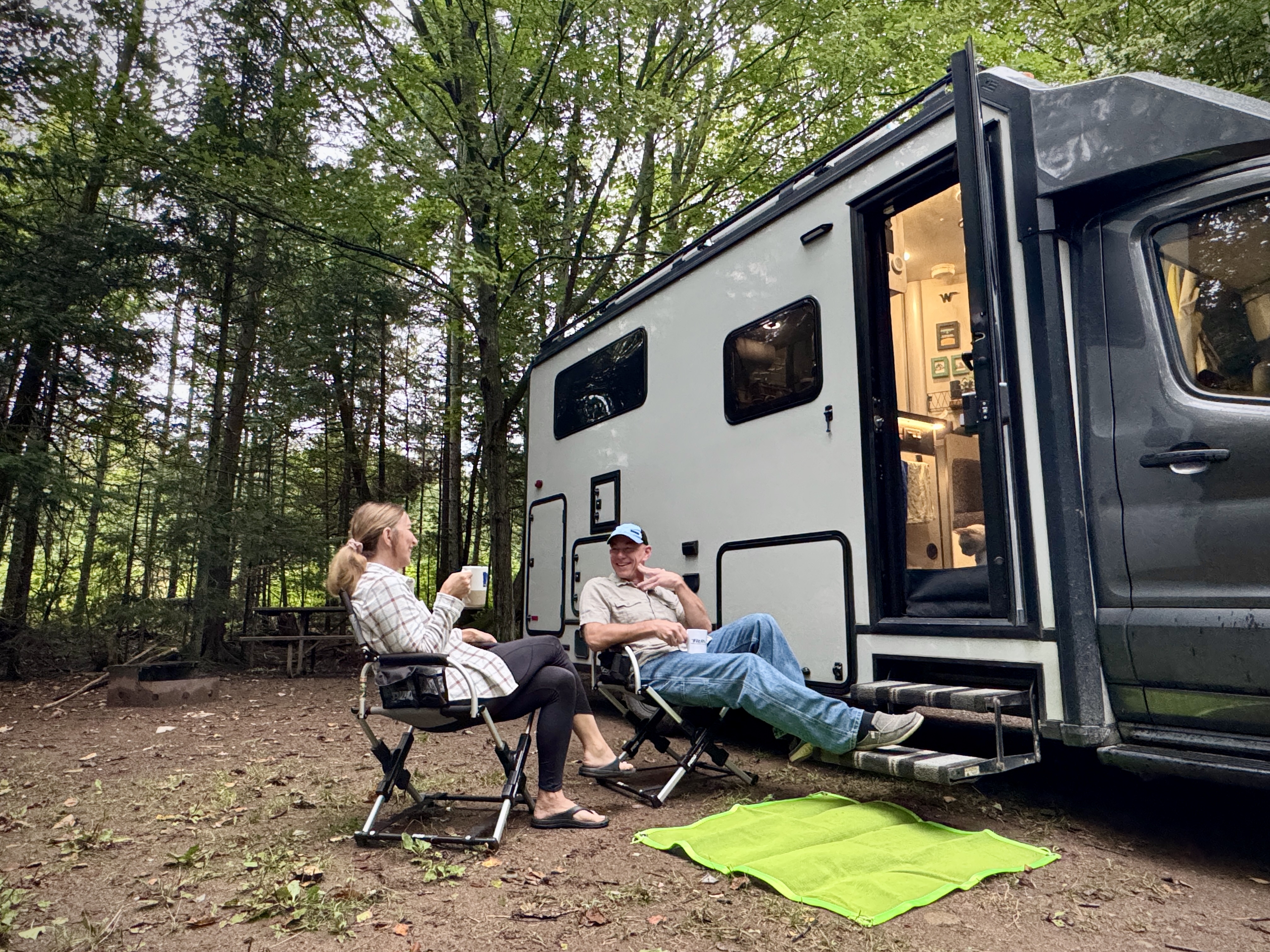A man and woman relax with their feet up next to their RV at a campsite surrounded by green forest at Pancake Bay Provincial Park. 